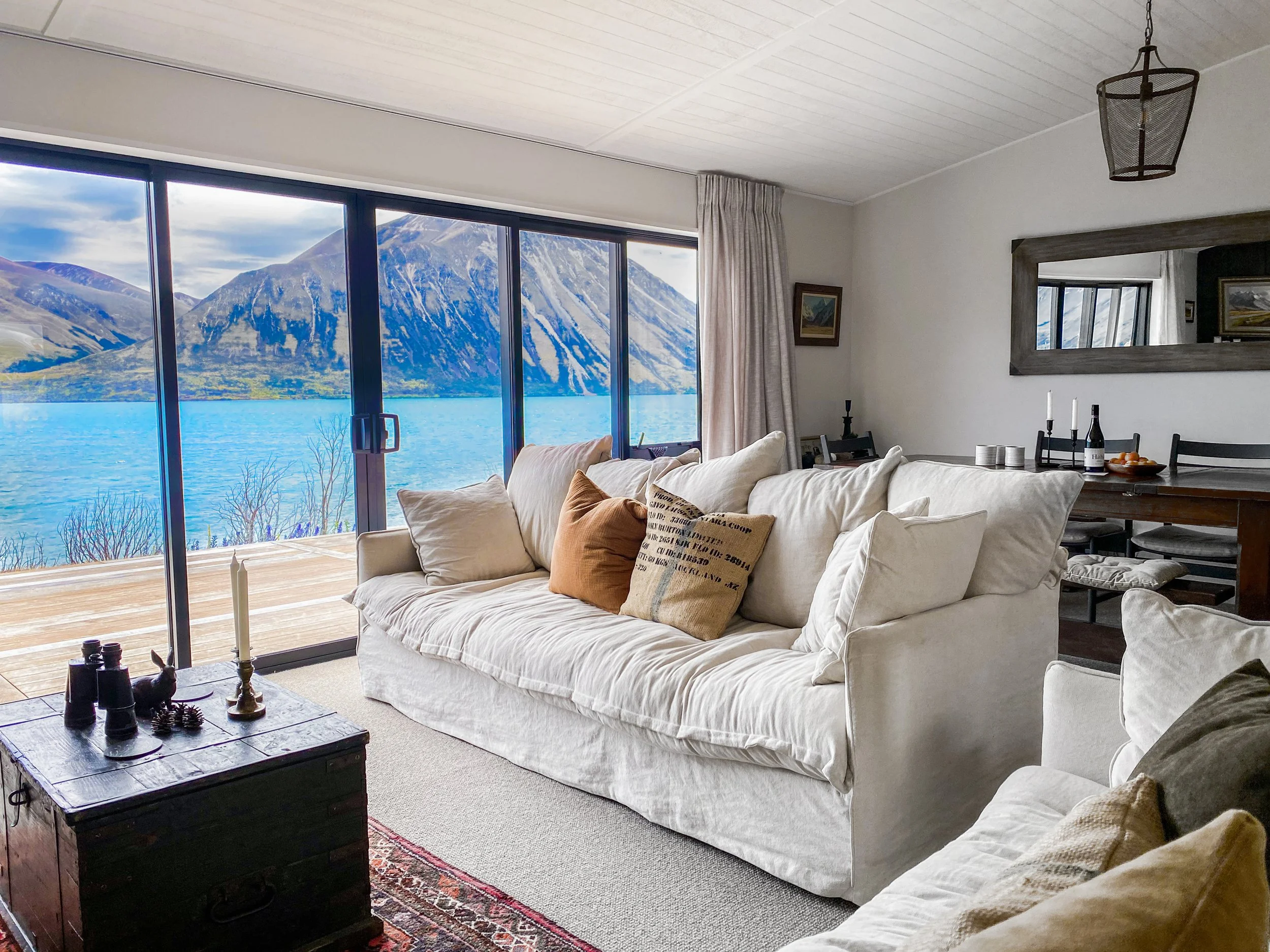 Living room with large glass sliding doors showcasing a mountain lake view, a beige sofa with throw pillows, a wooden coffee table with decorative items, and a wall mirror reflecting part of the room.