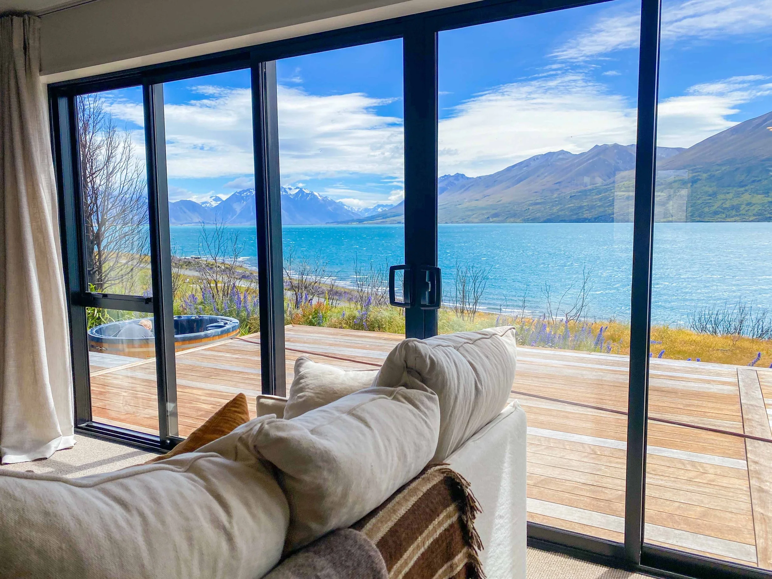 Living room with sliding glass door opening to a deck with a hot tub, overlooking a large body of water, with mountains in the distance and a partly cloudy sky. Lake Ohau Escape Lake-front AirBNB Accomodation, South Island, New Zealand.