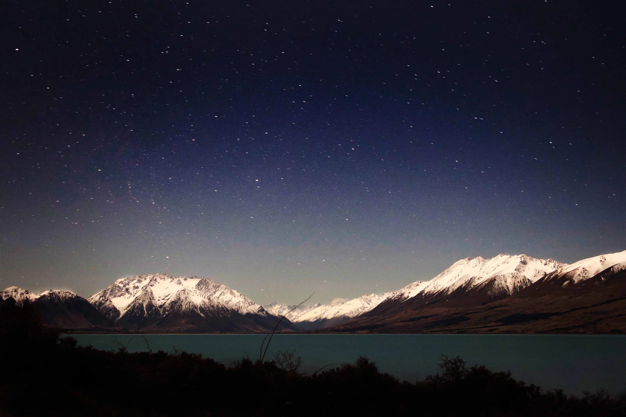 Night sky filled with stars above snow-capped mountains and a body of water in the foreground.