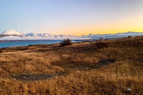 Open field with dry grass and a dirt path, mountains in the distance, and a cloudy sky at sunset. Twizel Ohau Canal Lake River Fishing Salmon Trout. Stay at Lake Ohau Escape AirBnb Accomodation.