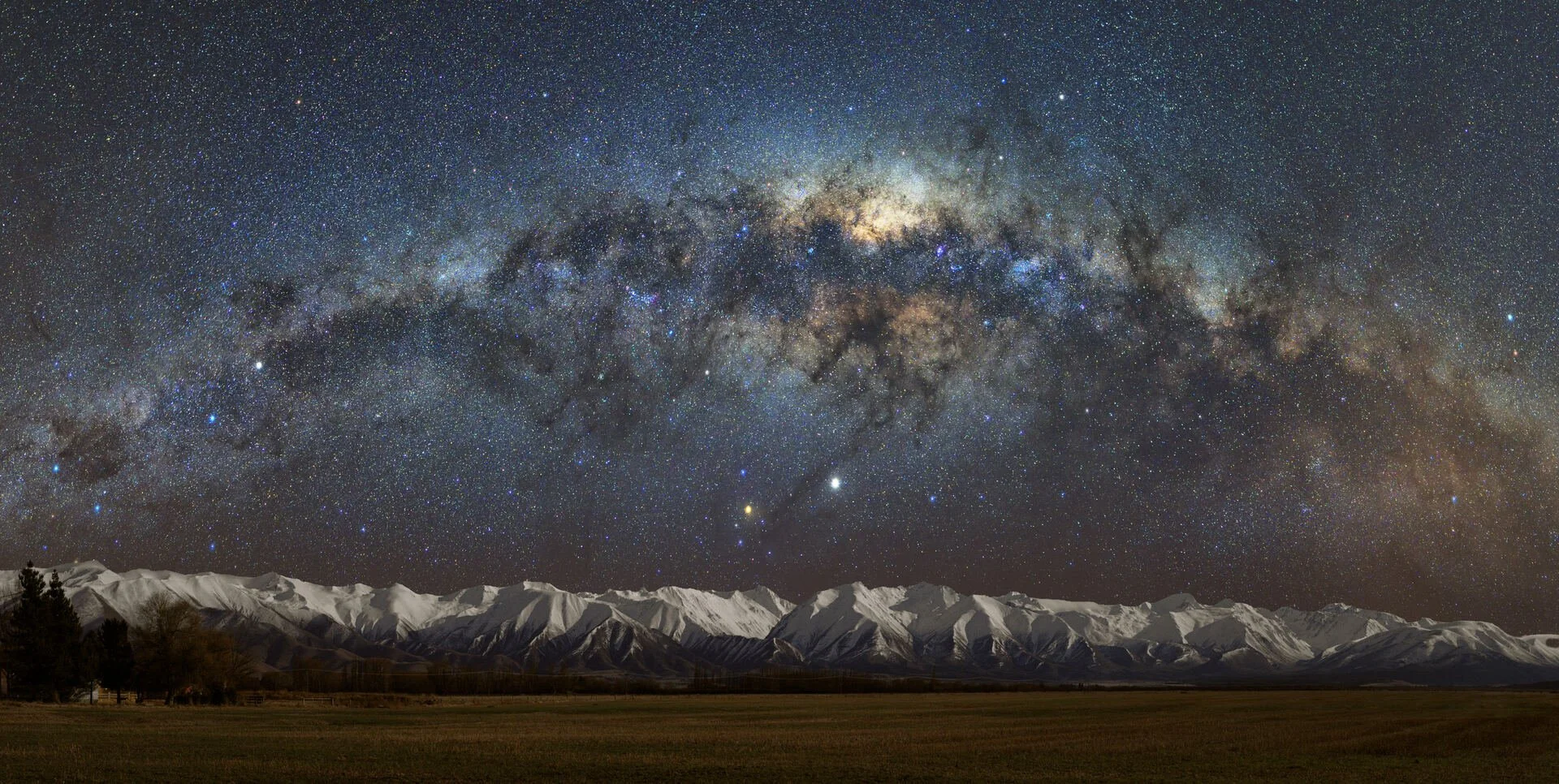 Night sky filled with stars and the Milky Way galaxy above snow-capped mountains and a grassy field. Lake Ohau Escape Lake-front AirBNB, South Island, New Zealand.