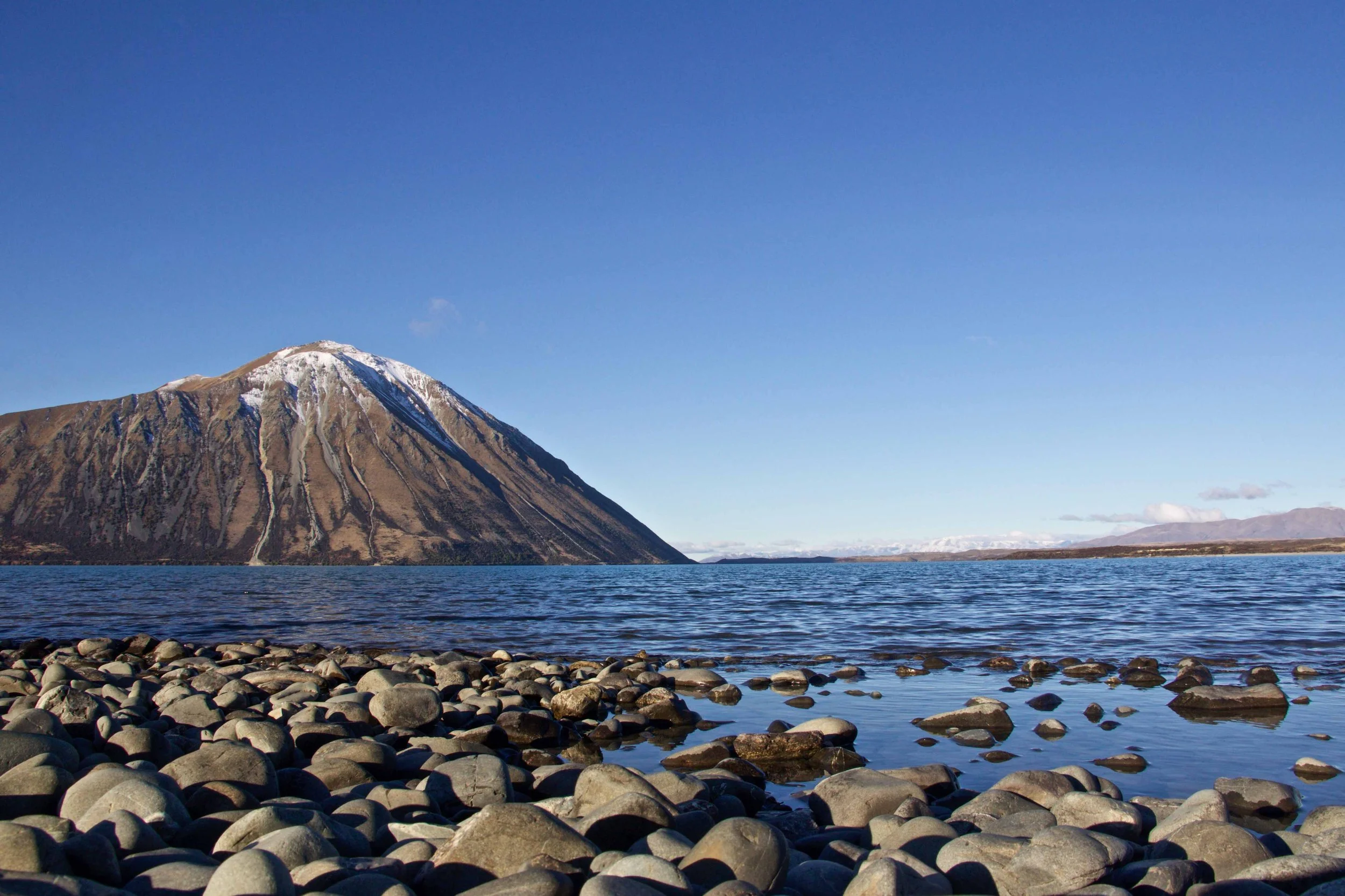 A mountain with snow at the peak rising steeply from a lake, with a rocky shoreline in the foreground and a clear blue sky above. Lake Ben Ohau Escape Lake-front AirBNB Accomodation, South Island, New Zealand.