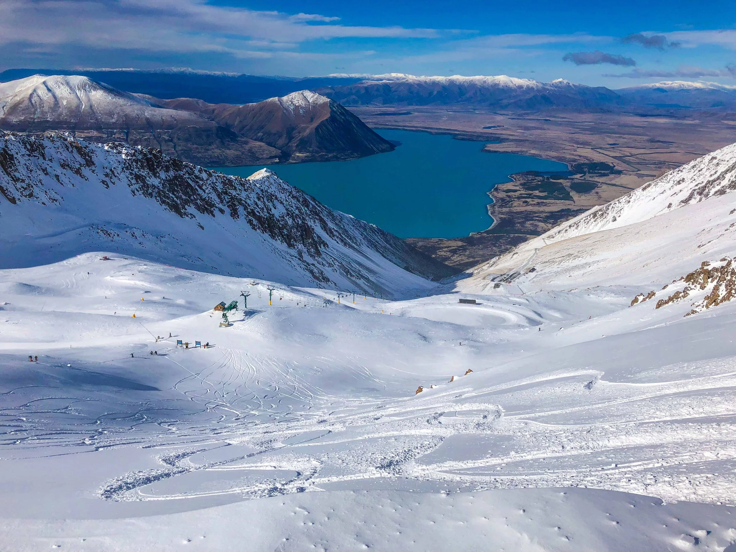 Snow-covered ski slope with ski lifts and skiers, mountains in the background, and a lake at the valley. Lake Ohau Escape Lake-front AirBNB Accomodation, South Island, New Zealand.