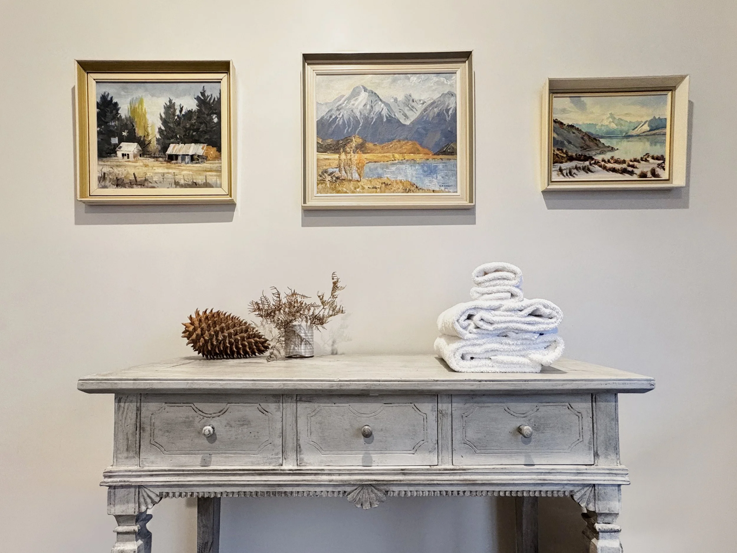 Display of three landscape paintings hung on a white wall, with a rustic wooden table below. The table has a stack of folded white towels and decorative natural elements.