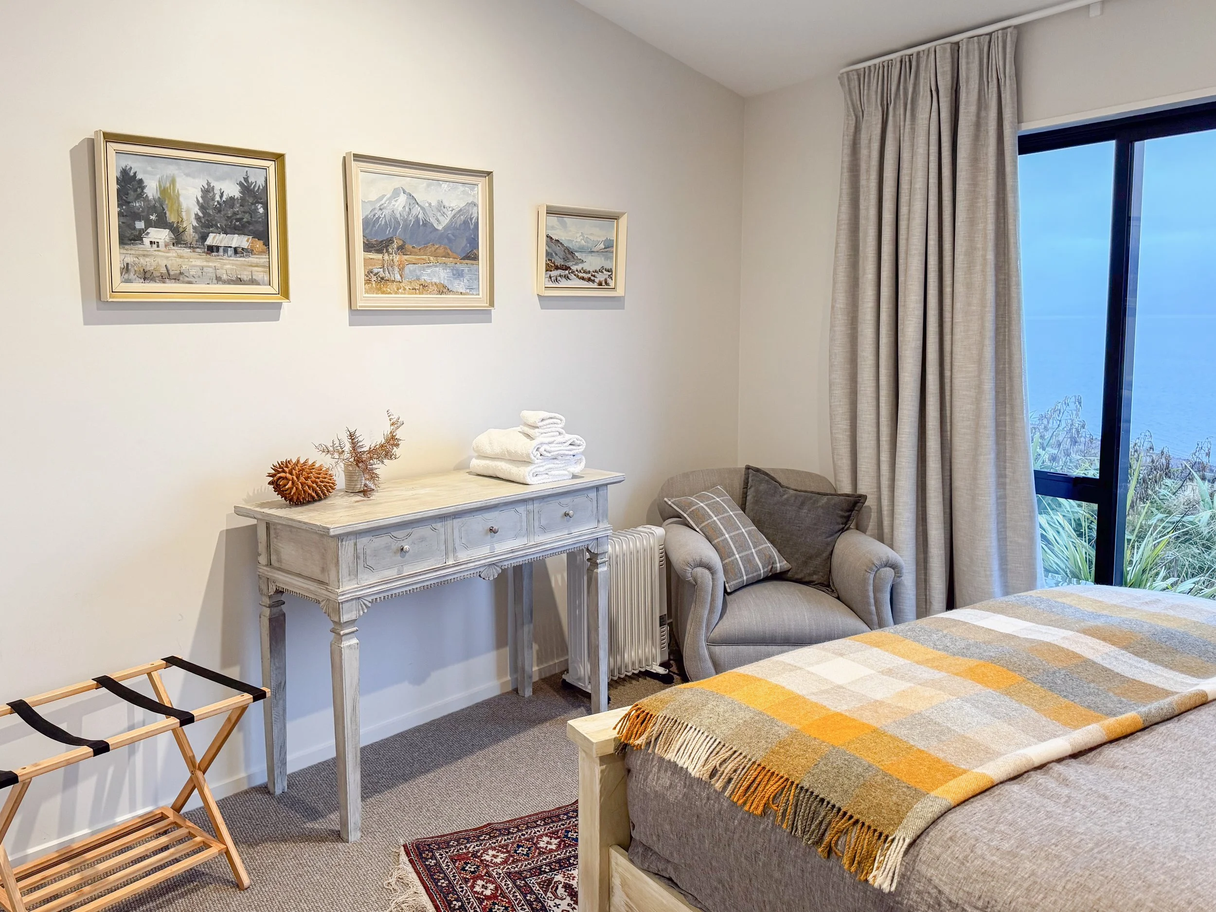 A cozy bedroom featuring a bed with a plaid blanket, a beige armchair with a pillow, a wooden dresser with folded towels and decorative items, artwork depicting mountains and nature on the wall, a window with beige curtains, and a small rug on the ca