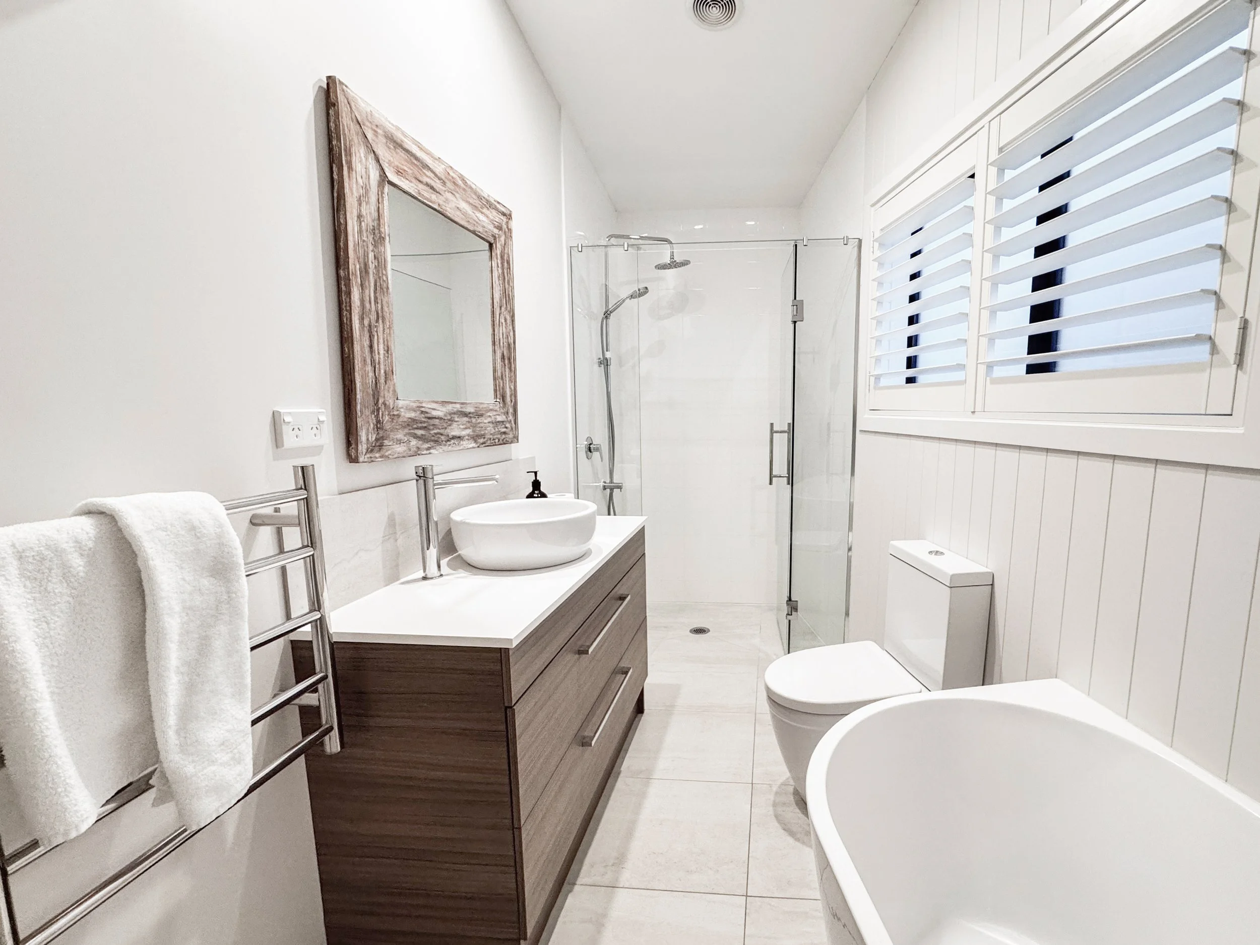 Modern bathroom with white walls, a wooden framed mirror, a towel rack with white towels, a vanity with a vessel sink, a glass-enclosed shower, a window with white shutters, and a toilet.