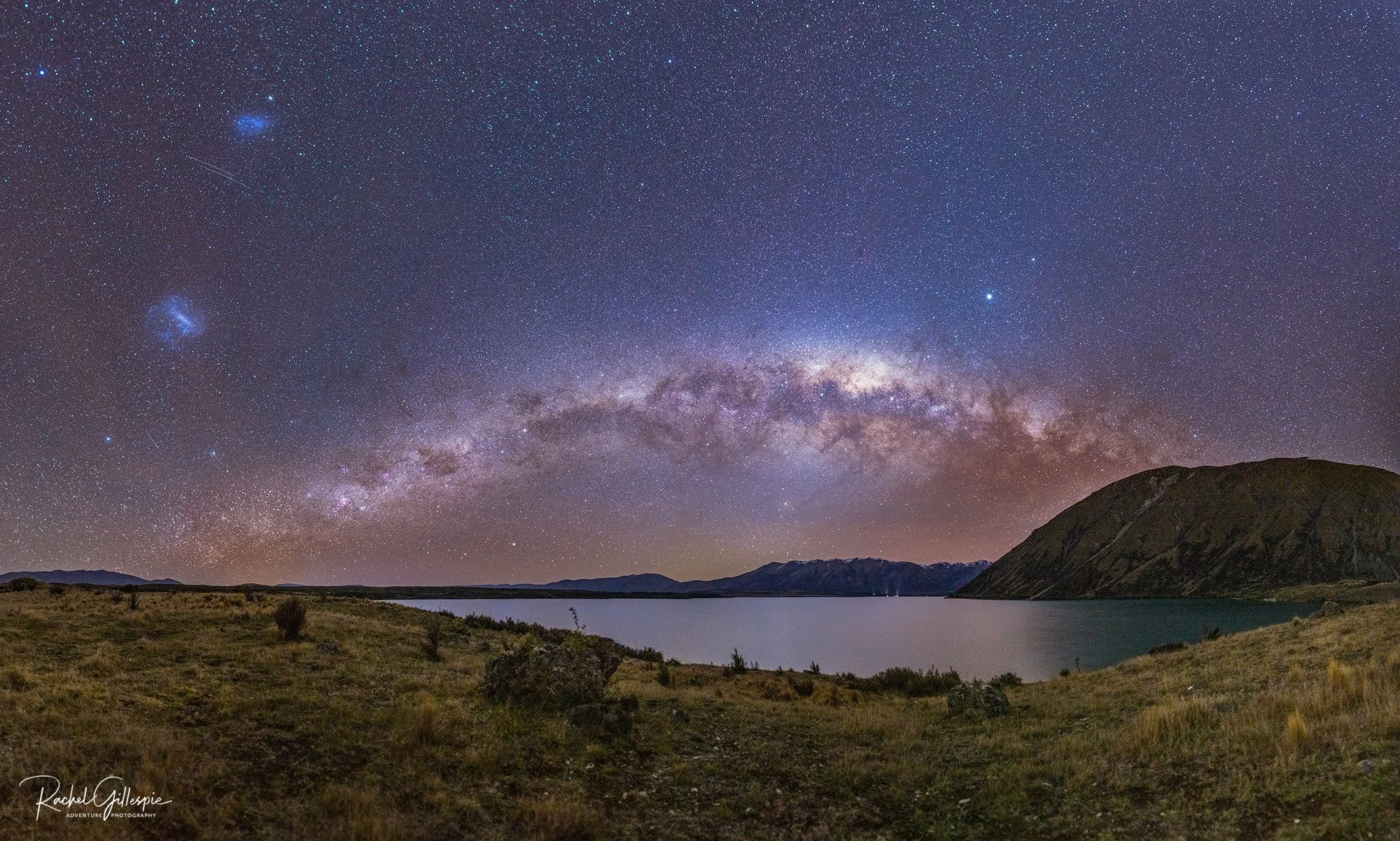 Night sky filled with stars and the Milky Way galaxy over a landscape with a lake, hills, and mountains. Lake Ohau Escape Lake-front AirBNB, South Island, New Zealand.