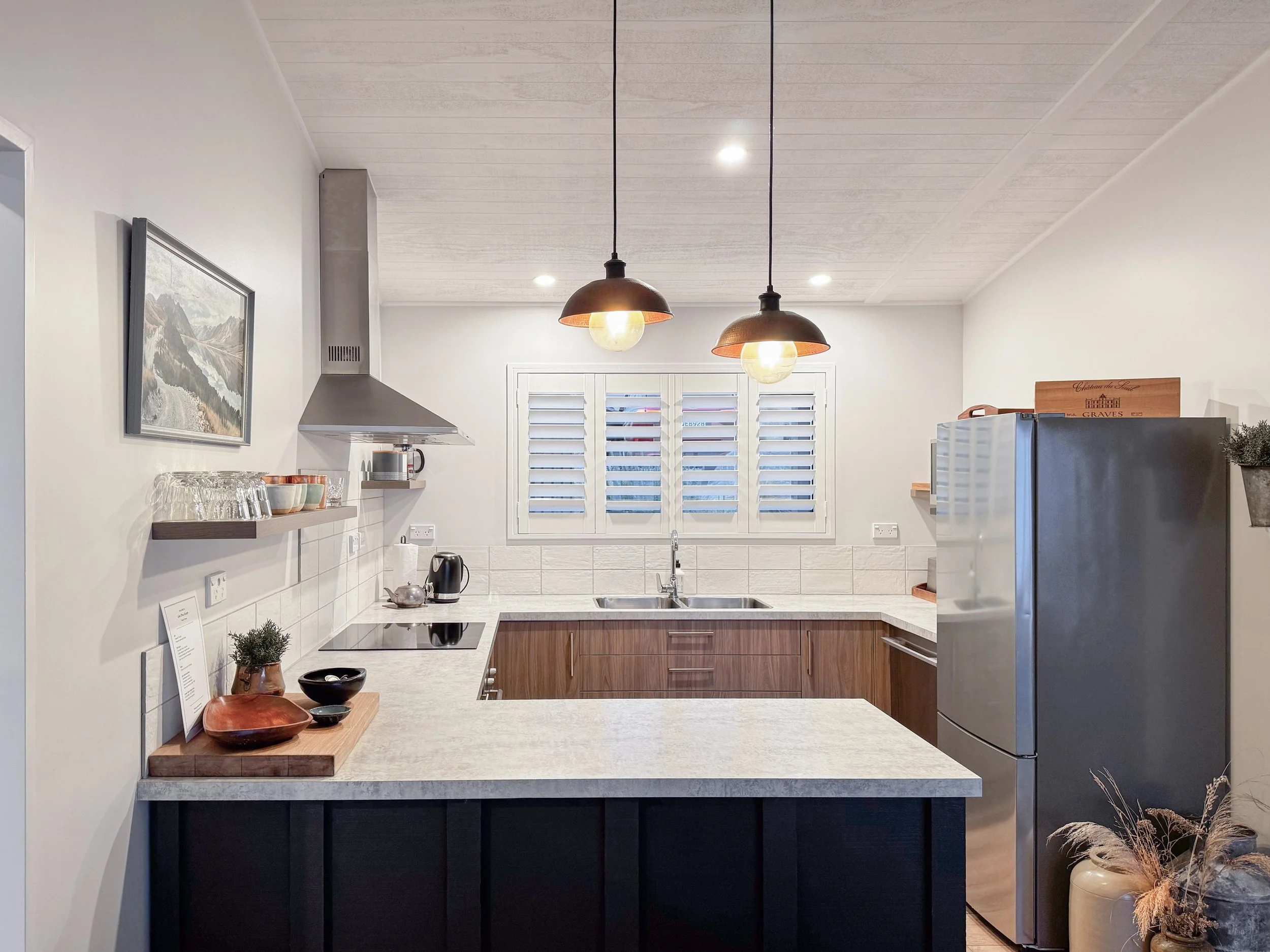 Modern kitchen with white walls, wooden cabinets, two black pendant lights, a window with white shutters, and various kitchen items like bowls, a kettle, and decor.