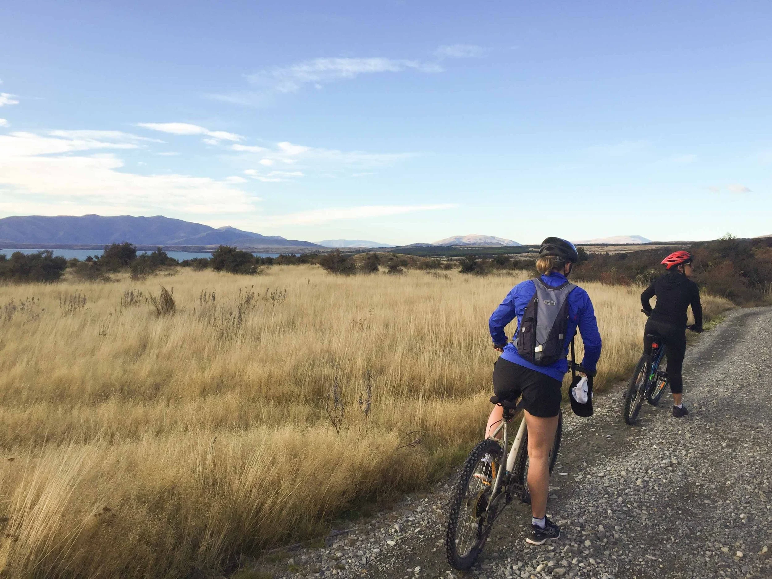 Two people cycling on a gravel trail through a grassy field with mountains and blue sky in the background.