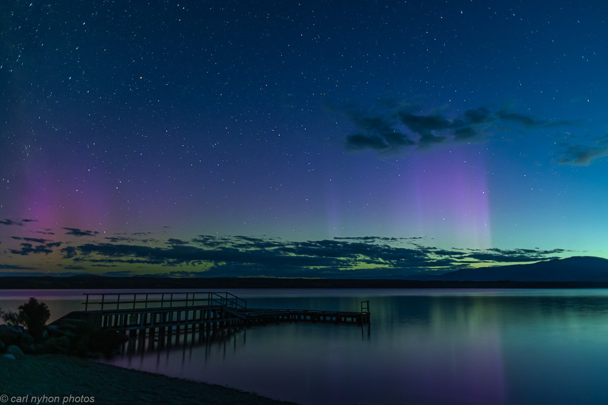 Nighttime scene of teal waters with a small dock extending into a calm lake, under a starry sky with visible aurora borealis and dark clouds. Lake Ohau Escape Lake-front AirBNB Accomodation, South Island, New Zealand.