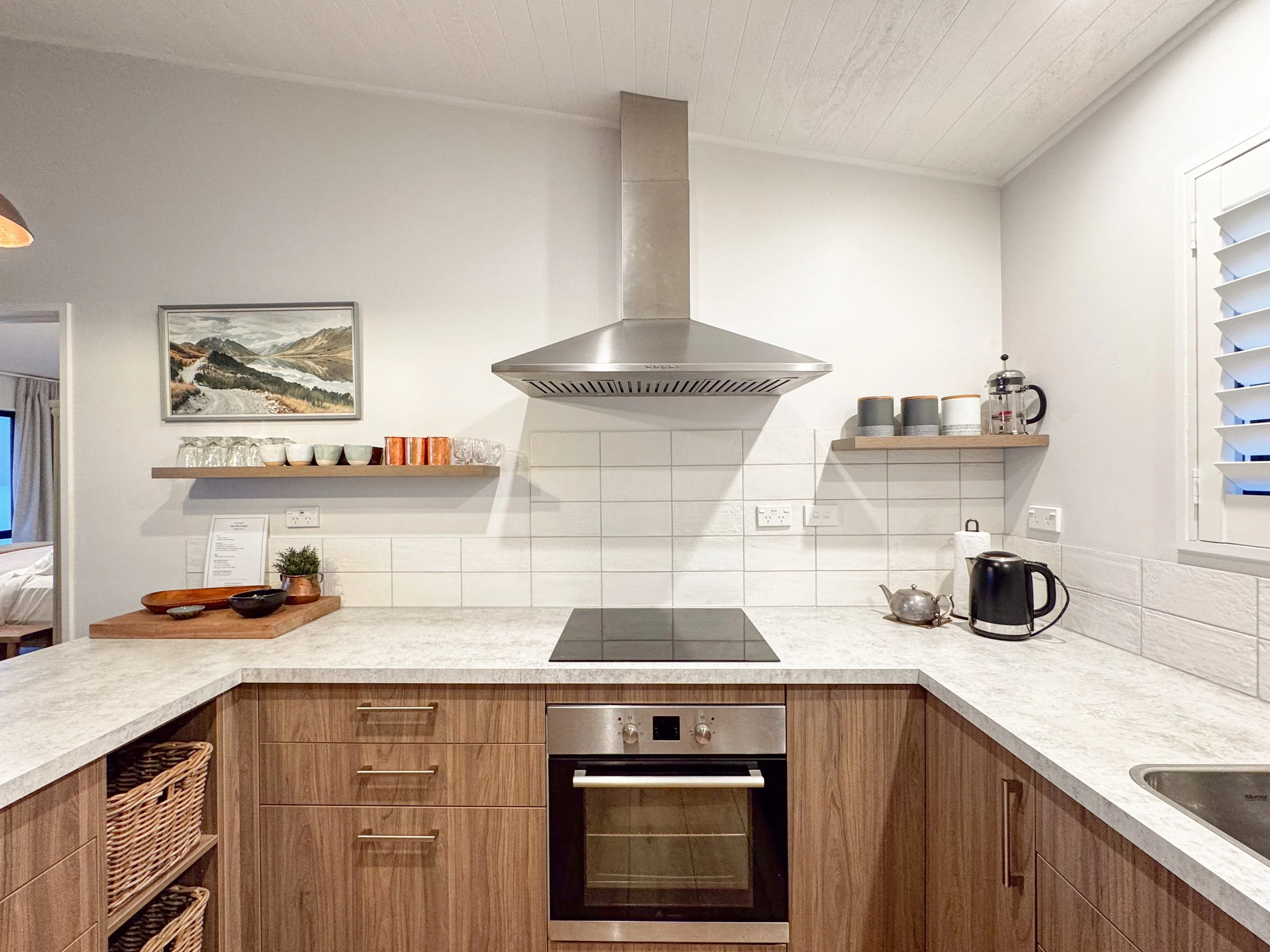 Modern kitchen with white walls, wooden cabinetry, a marble countertop, a stainless steel oven, a range hood, and small appliances on a floating shelf, including a French press, cups, and a kettle.