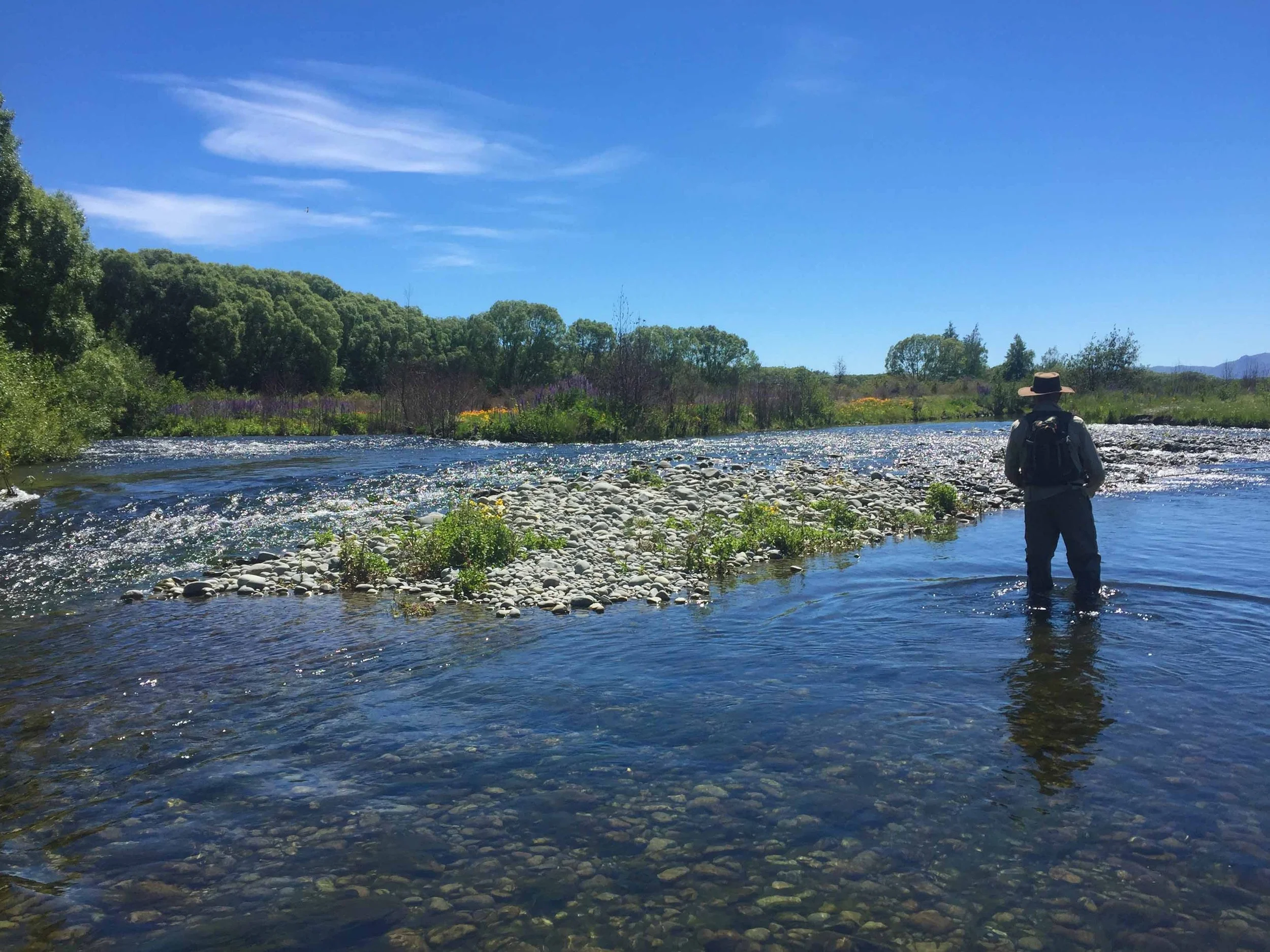 A person wearing a hat and backpack standing by a river with a forested area and blue sky in the background.