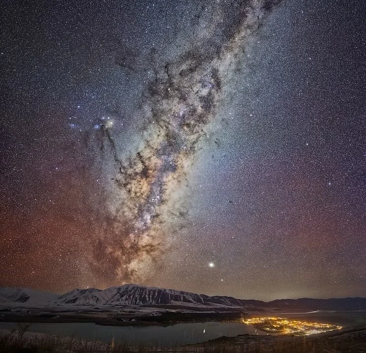 Night sky filled with stars and the Milky Way galaxy above snow-capped mountains, a body of water, and a small town with illuminated buildings. Lake Ohau Escape Lake-front AirBNB, South Island, New Zealand.