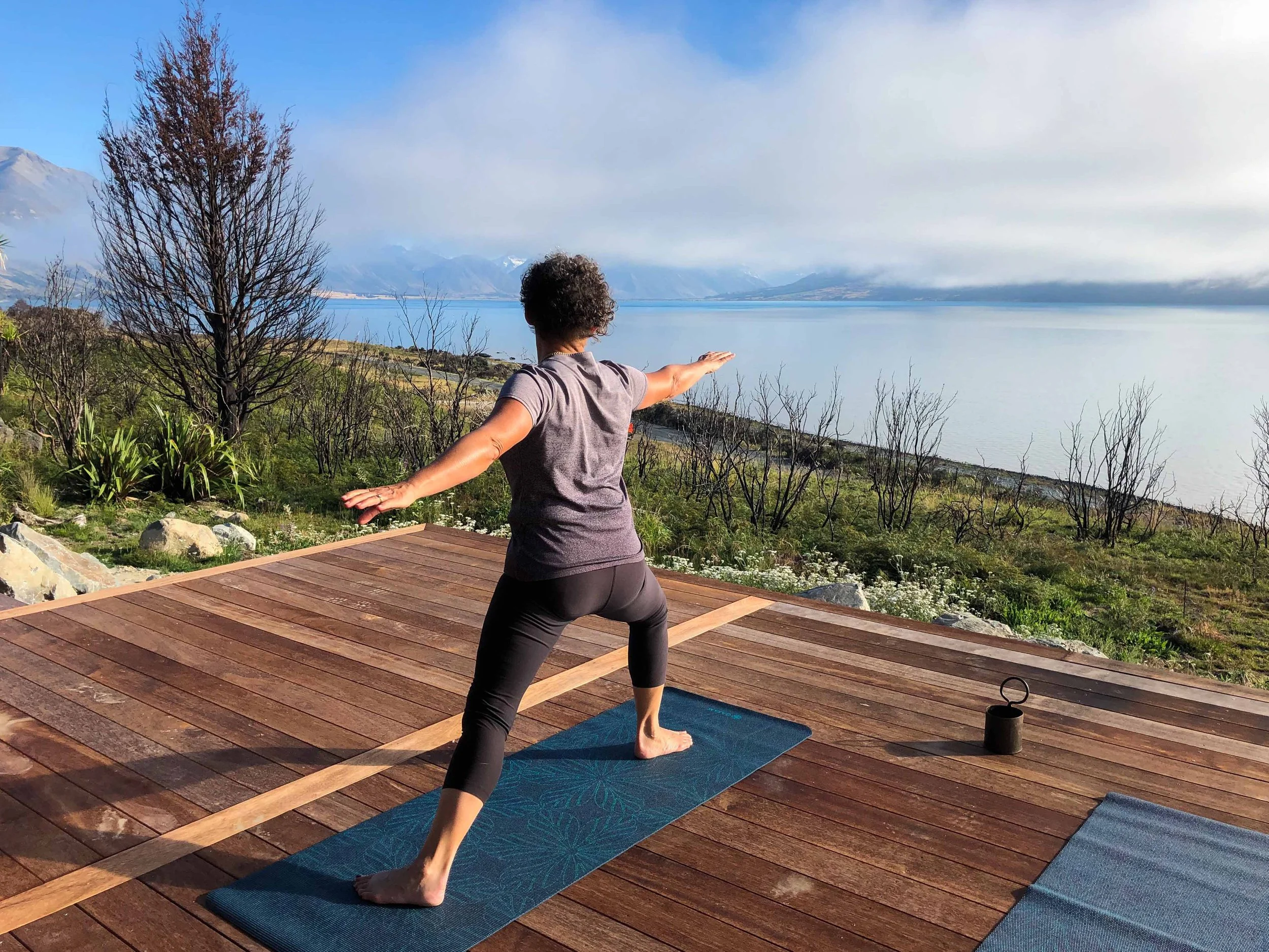 Person practicing yoga outdoors on a wooden deck overlooking a lake with mountains in the background.