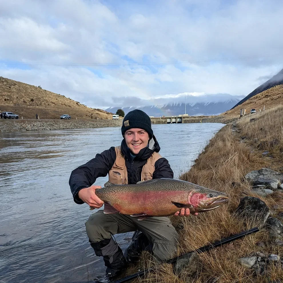 A person smiling and holding a large rainbow trout fish near a river with hills and mountains in the background.