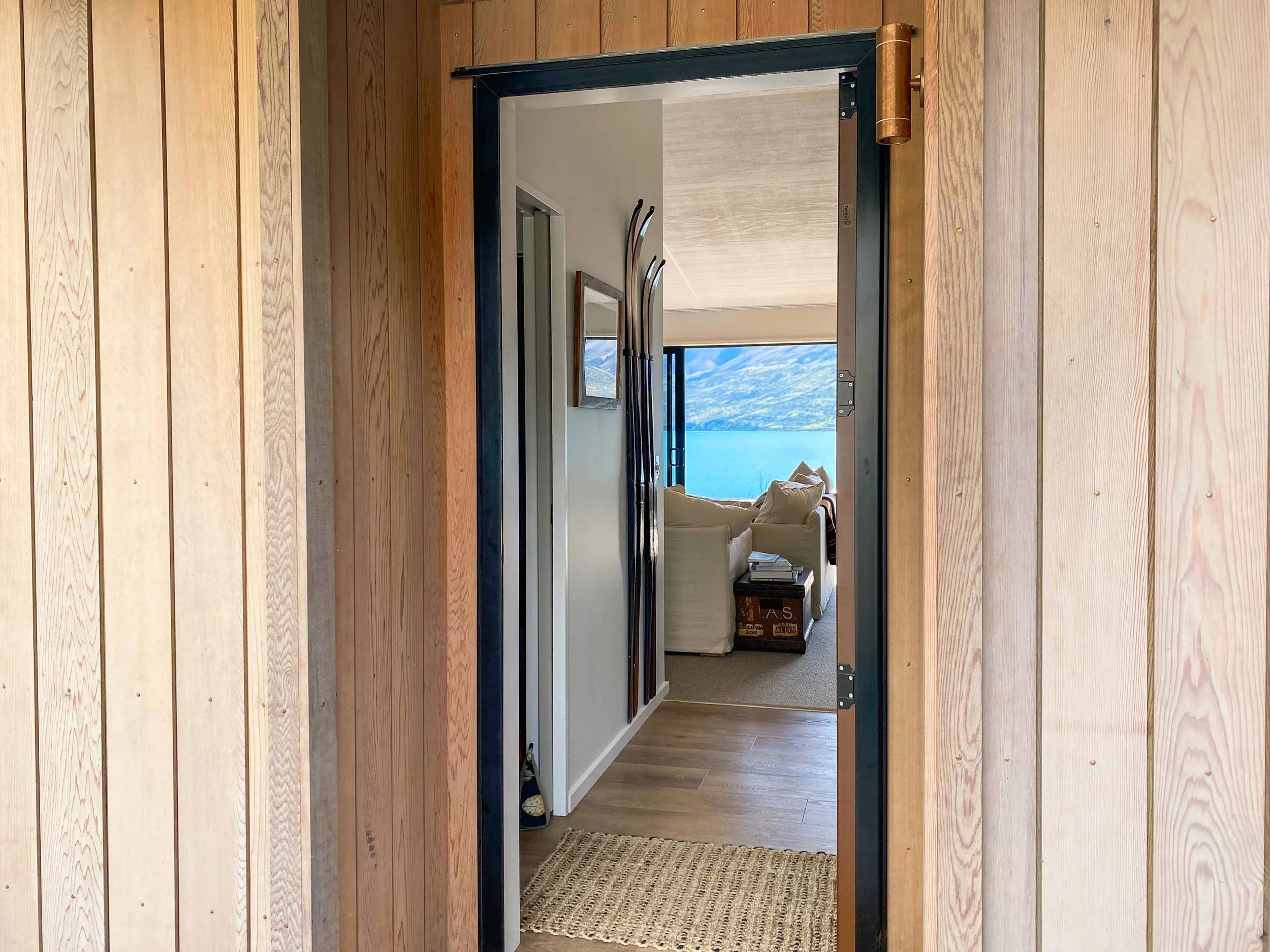 View through an open doorway into a living room with a large window showing a lake and mountains in the background. The room has beige walls and a beige couch, with some books and decor on a small table beside it.