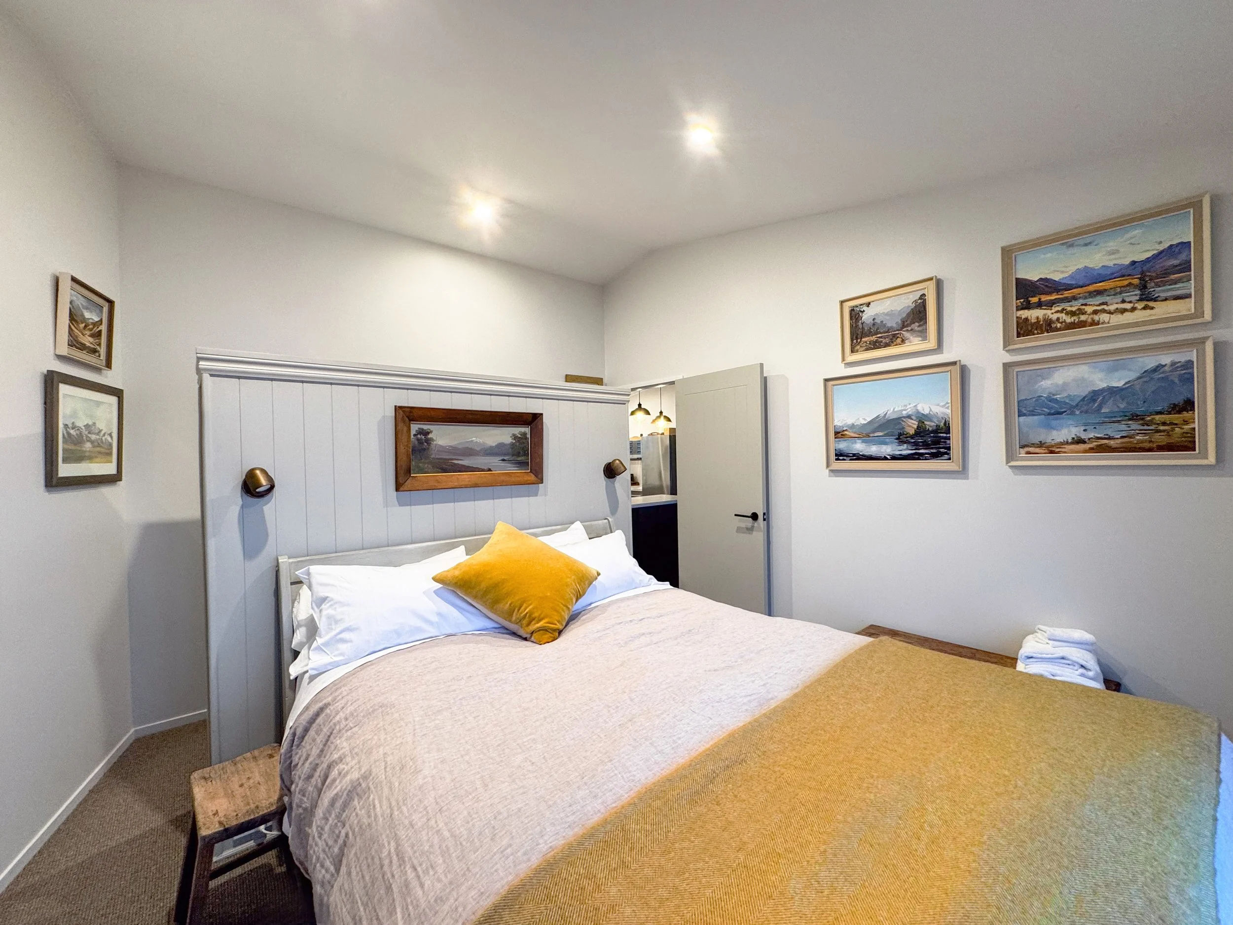 A cozy bedroom with a large bed covered in beige and white bedding, decorated with a yellow pillow. The wall behind the bed has framed landscape paintings mounted on it, and there are two black wall lamps on either side of the bed. The room has white