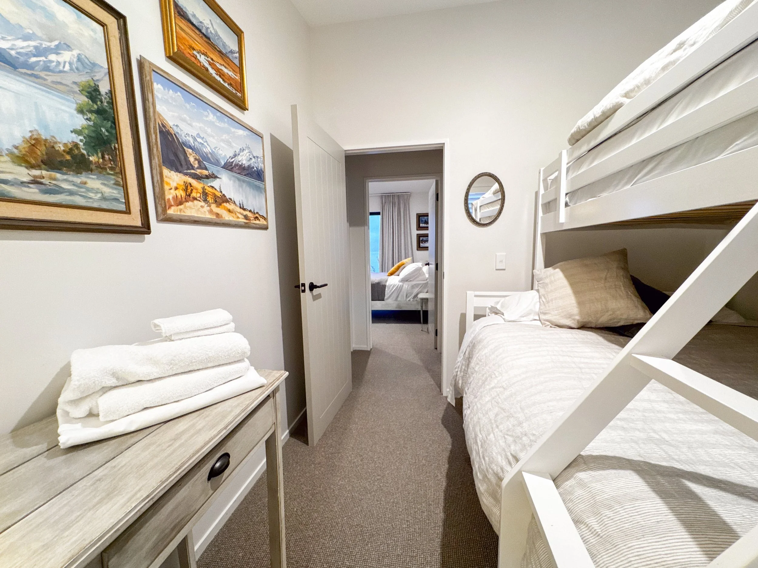 A bedroom with a bunk bed on the right, a dresser with folded towels on the left, and a mirror on the wall. There are framed landscape paintings on the wall, and another bedroom visible through the open door.
