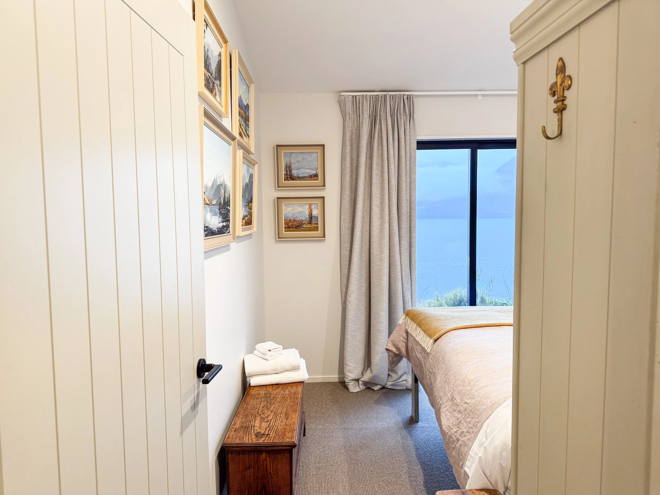 A cozy bedroom with a large window overlooking a body of water and mountains. The room has beige walls, framed landscape paintings, a bed with a tan quilt, and beige curtains. There is a wooden bench and a stack of white towels.
