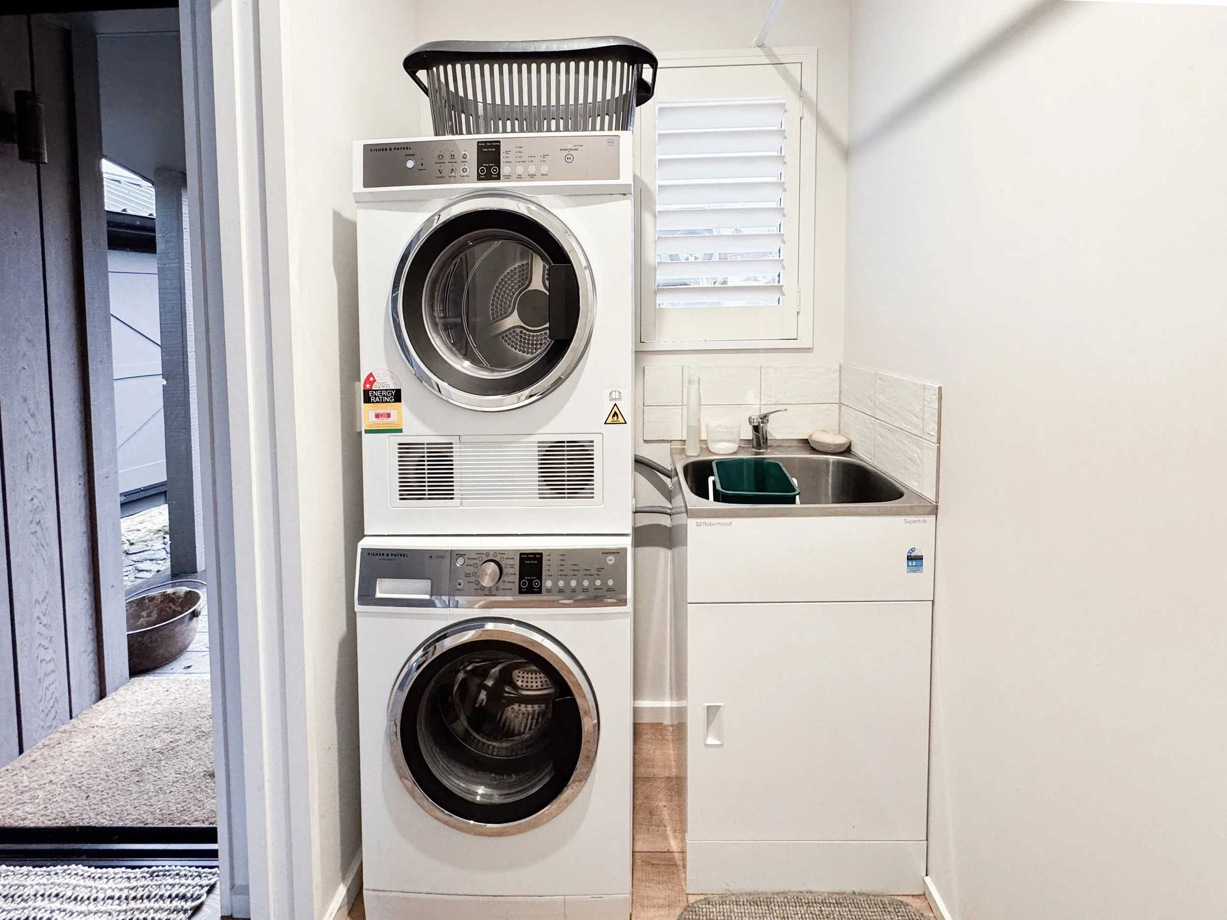 A laundry room with a stacked white washer and dryer. On top of the dryer is a black laundry basket. To the right, there is a small sink with a faucet, a soap dispenser, and a mirror above it. A white door with louvers is visible, and a small black w