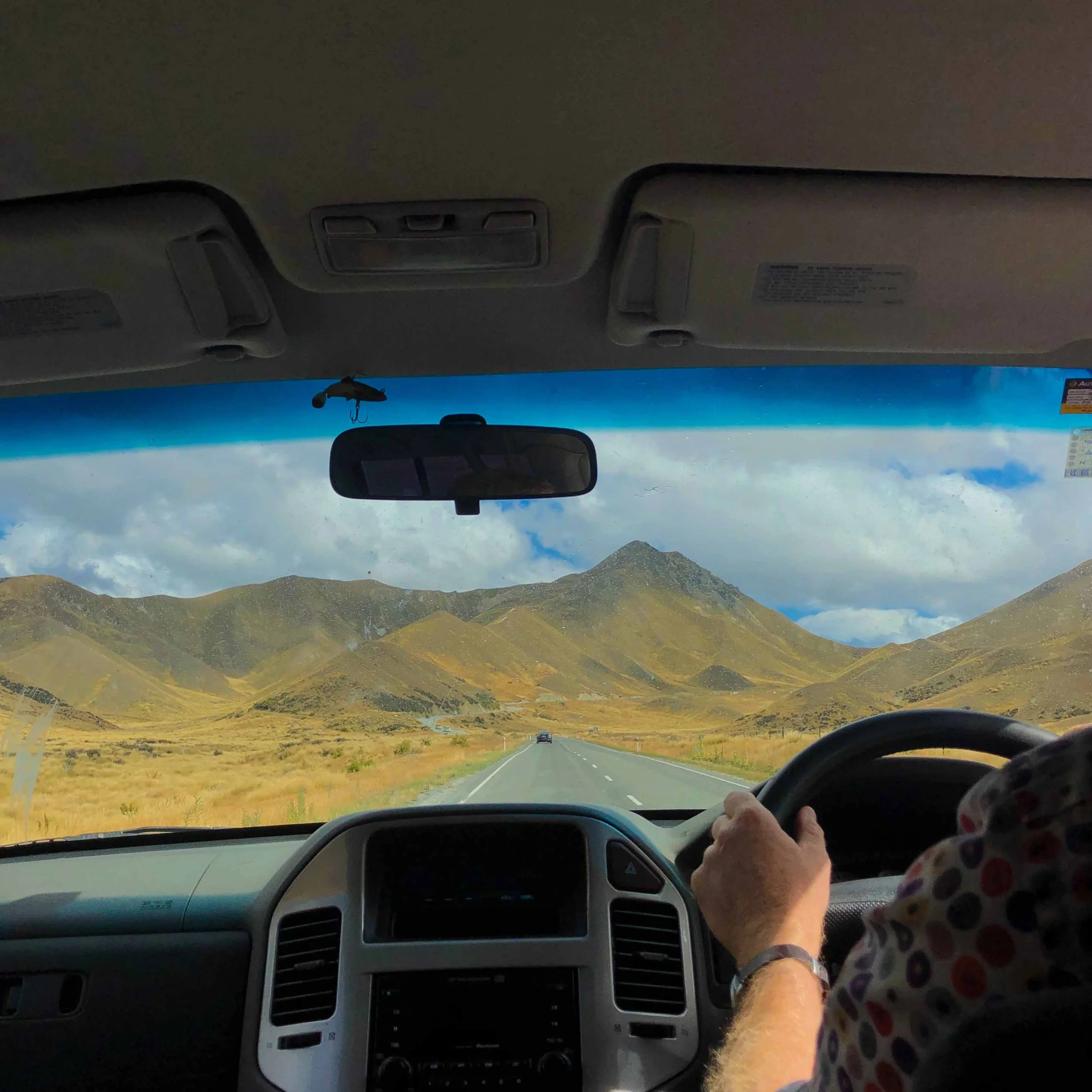 Inside a vehicle looking at the dashboard and windshield, with a mountainous landscape and cloudy sky visible outside.