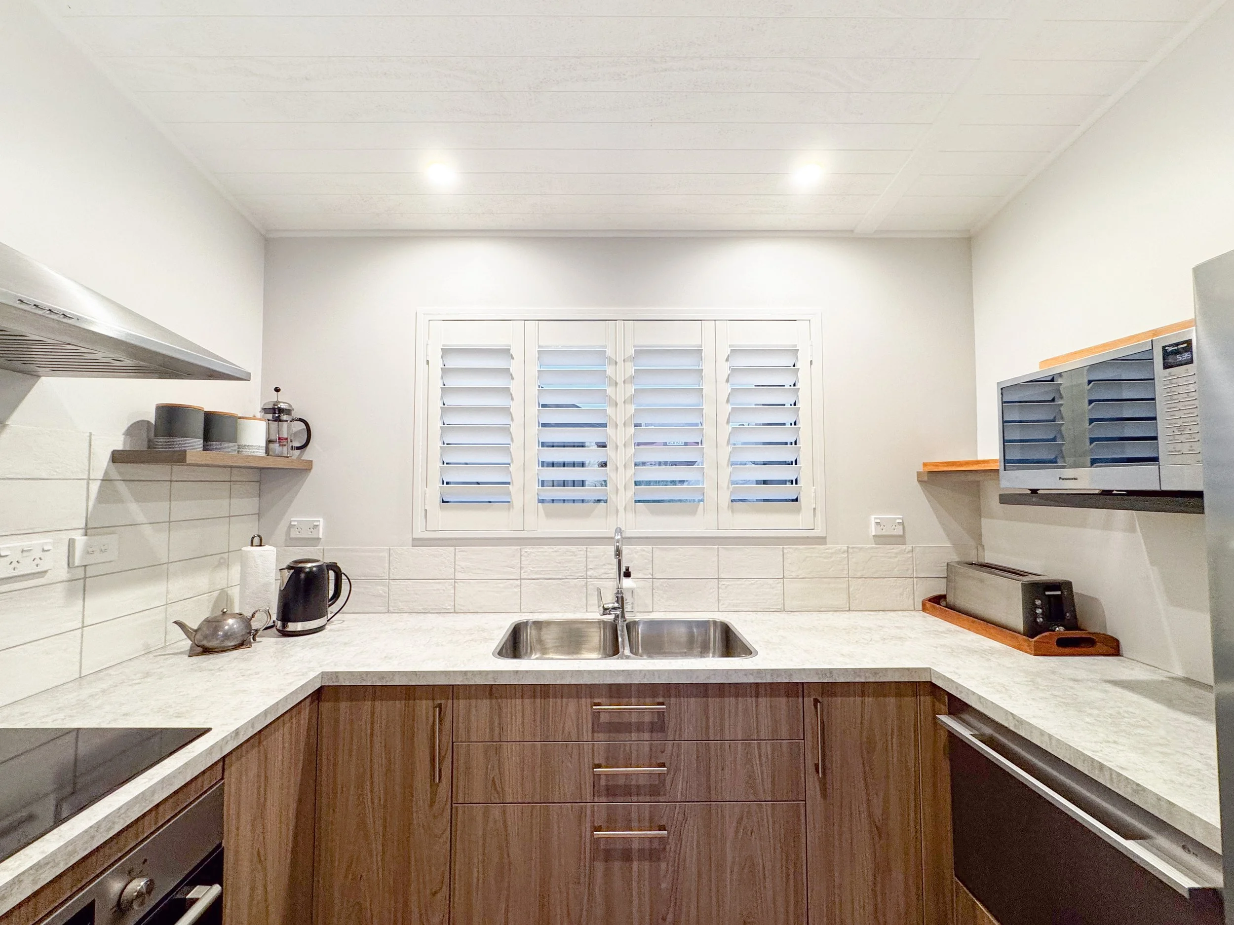 Kitchen with white cabinets, a window with shutters, and a granite countertop featuring a sink, kettle, microwave, and shelf with dishes.