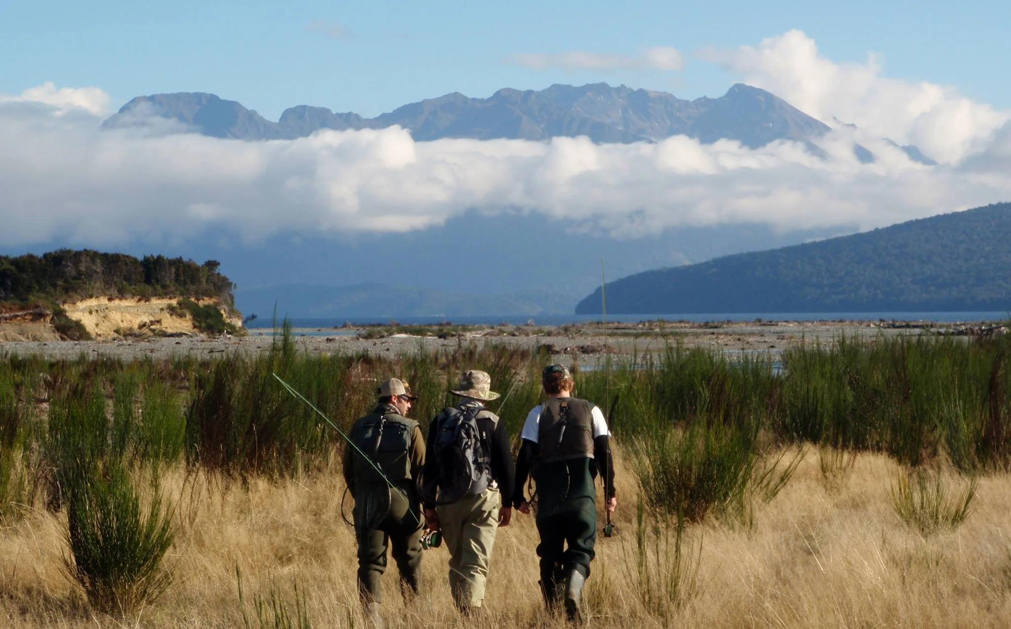 A group of hikers walking through grassy terrain near a body of water with islands and mountains in the background, under a partly cloudy sky. Twizel Ohau Canal Lake River Fishing Salmon Trout. Stay at Lake Ohau Escape AirBnb Accomodation.