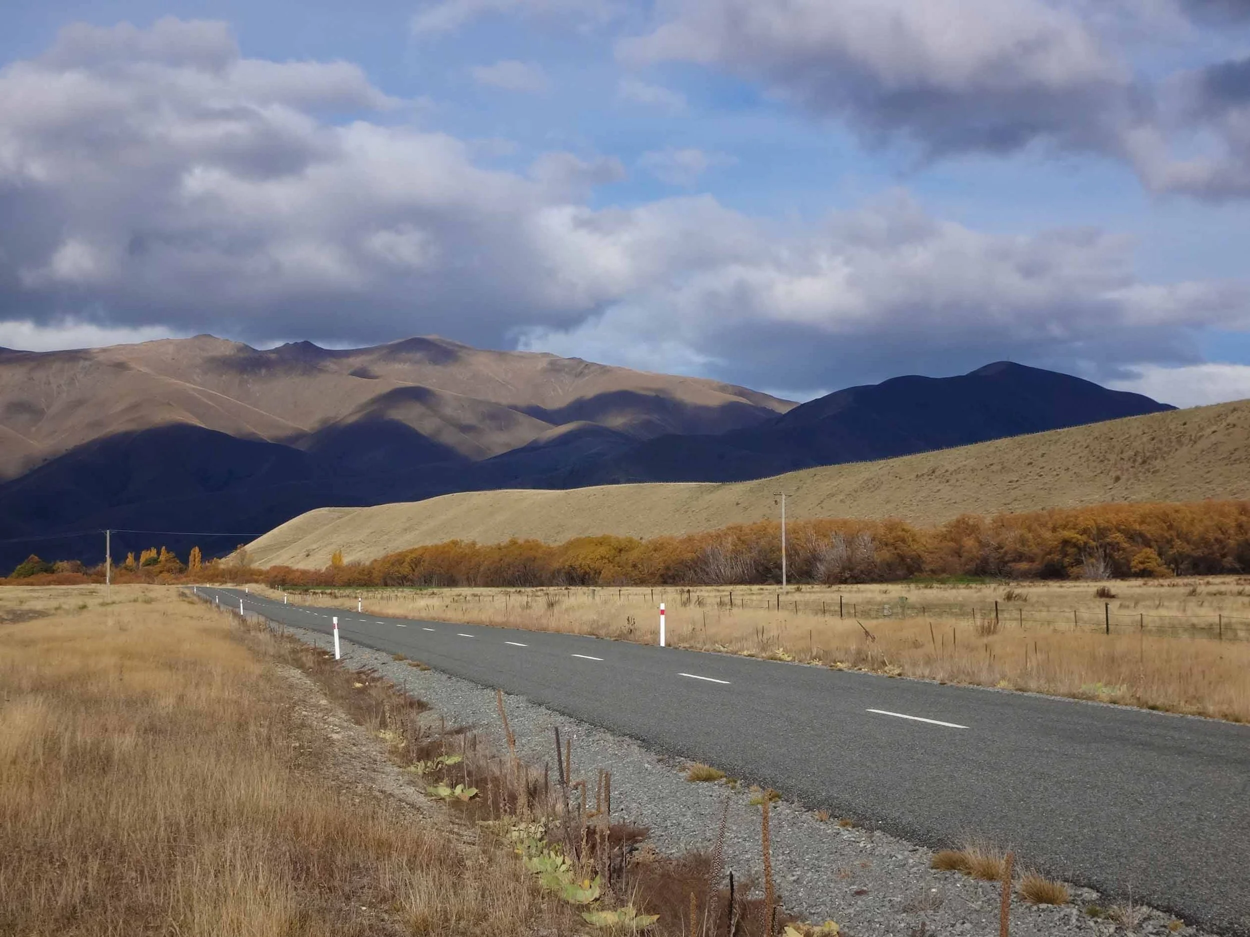 A rural road running through a landscape with grassy fields and hills in the background under a cloudy sky.
