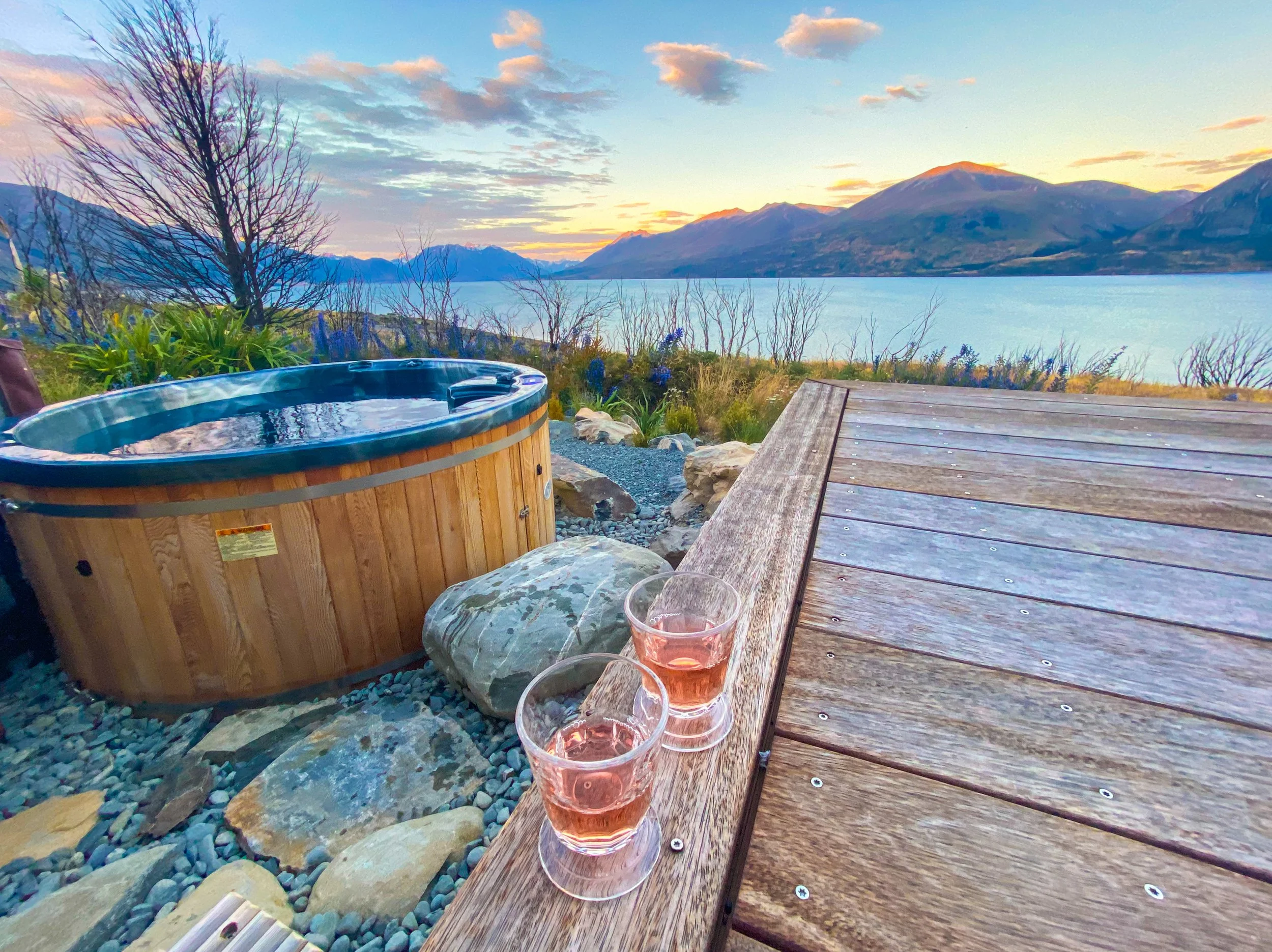 A lakeside outdoor setting during sunset with a hot tub, three glasses of pink drink on a wooden table, and mountains in the background.