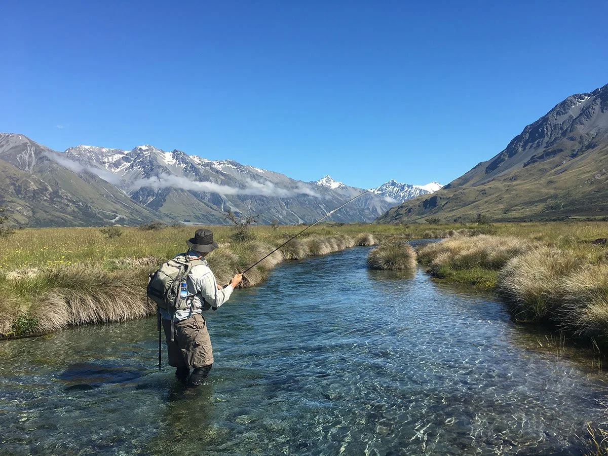 Person wearing outdoor gear fishing in a clear mountain stream with snow-capped mountains in the background. Twizel Ohau Canal Lake River Fishing Salmon Trout. Stay at Lake Ohau Escape AirBnb Accomodation.