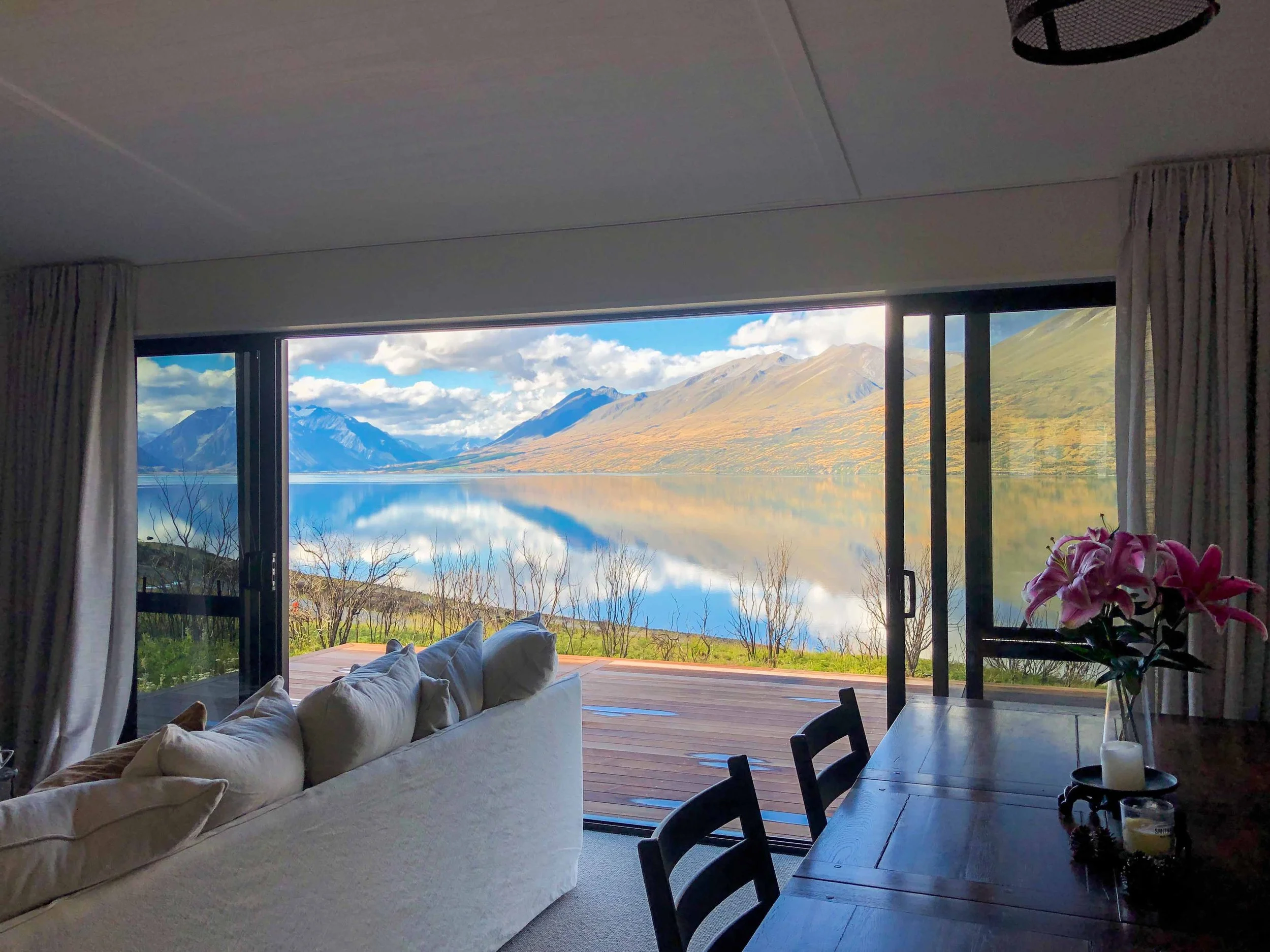 Living room with large open sliding glass doors revealing mountain landscape and lake outside. White sofa with pillows on the left, dining table and chairs on the right, and pink flowers on the table. Lake Ohau Escape Lake-front AirBNB Accomodation, 