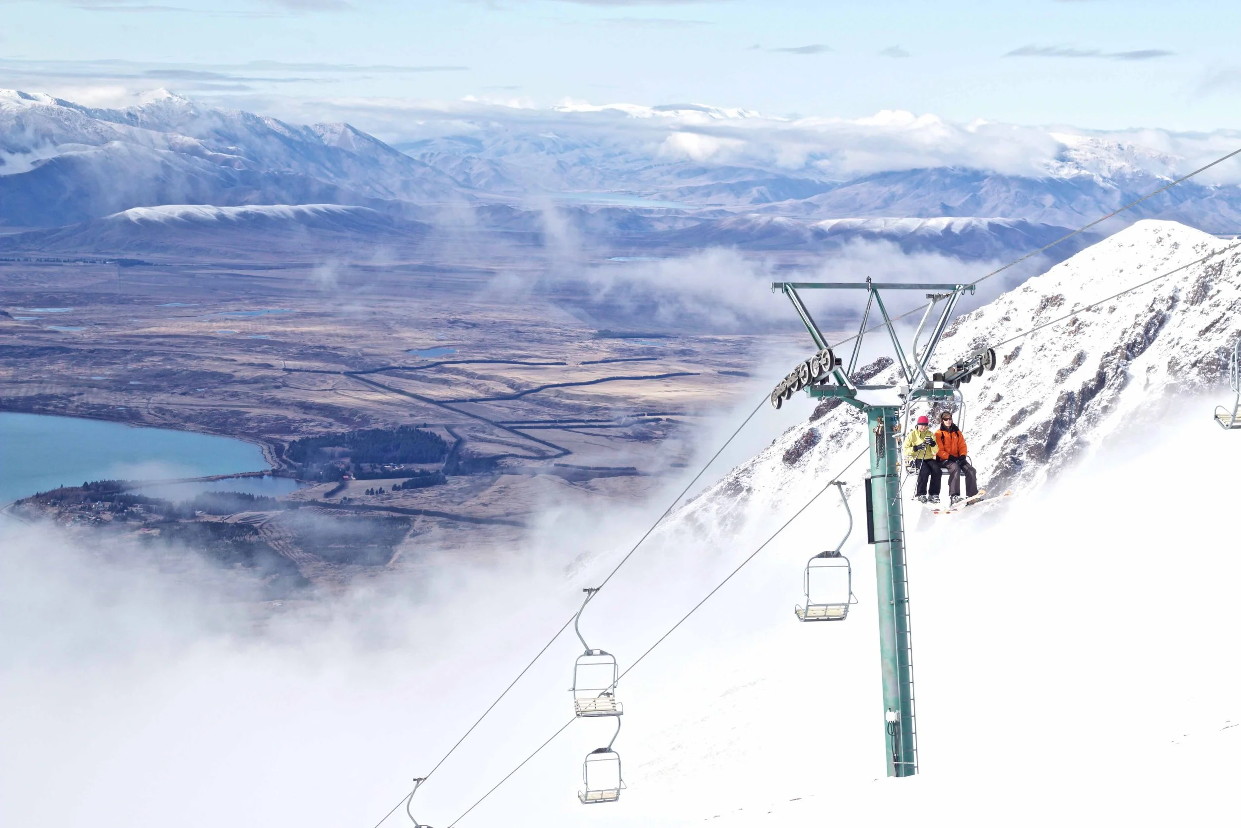 Skiers riding a ski lift in a snowy mountain landscape with snow-capped peaks, clouds, and a valley below.
