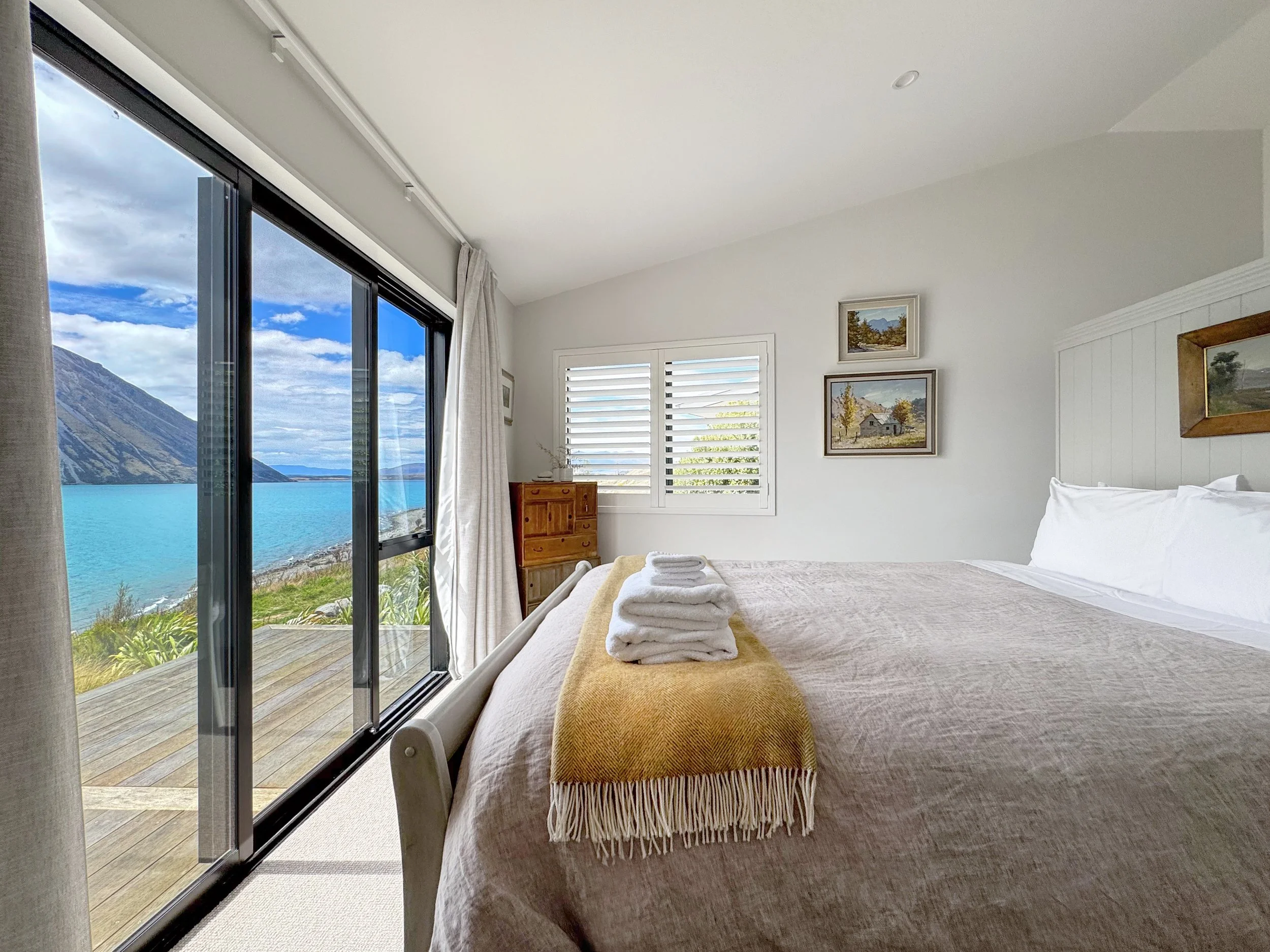 A bedroom with a large sliding glass door opening to a balcony overlooking a body of water and mountains. There is a bed with white bedding, stacked towels on the bed, framed landscape paintings on the wall, and white plantation shutters on another window.