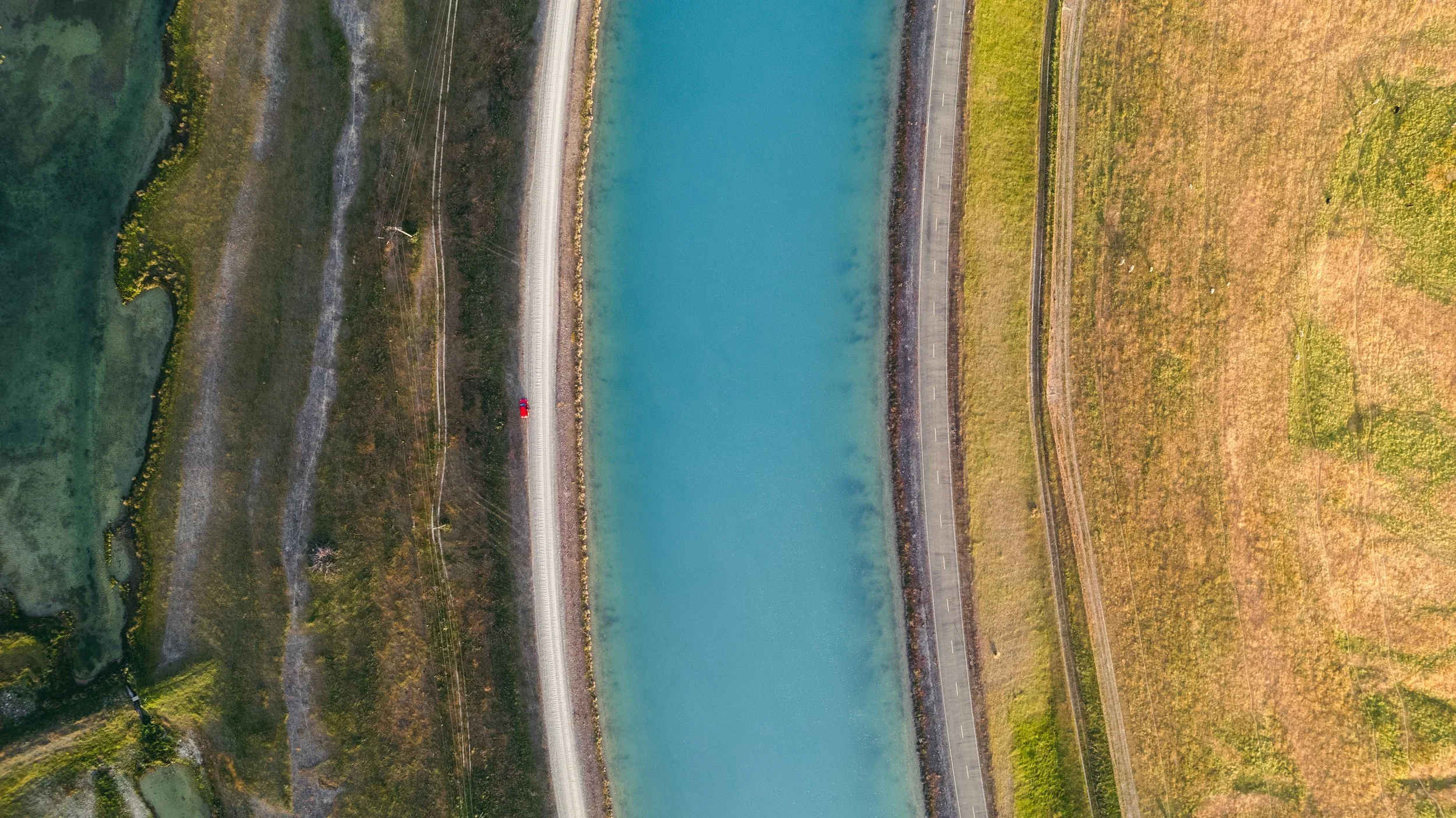 An aerial view of a reservoir with blue water, surrounded by a road and grassy fields. Twizel Ohau Canal Lake River Fishing Salmon Trout. Stay at Lake Ohau Escape AirBnb Accomodation.