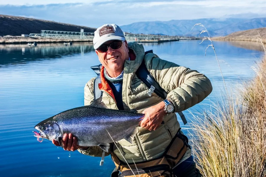 Smiling man holding a large fish by a river, wearing sunglasses, a beige jacket, cap, and watch, with mountains in the background.Twizel Ohau Canal Lake River Fishing Salmon Trout. Stay at Lake Ohau Escape AirBnb Accomodation.