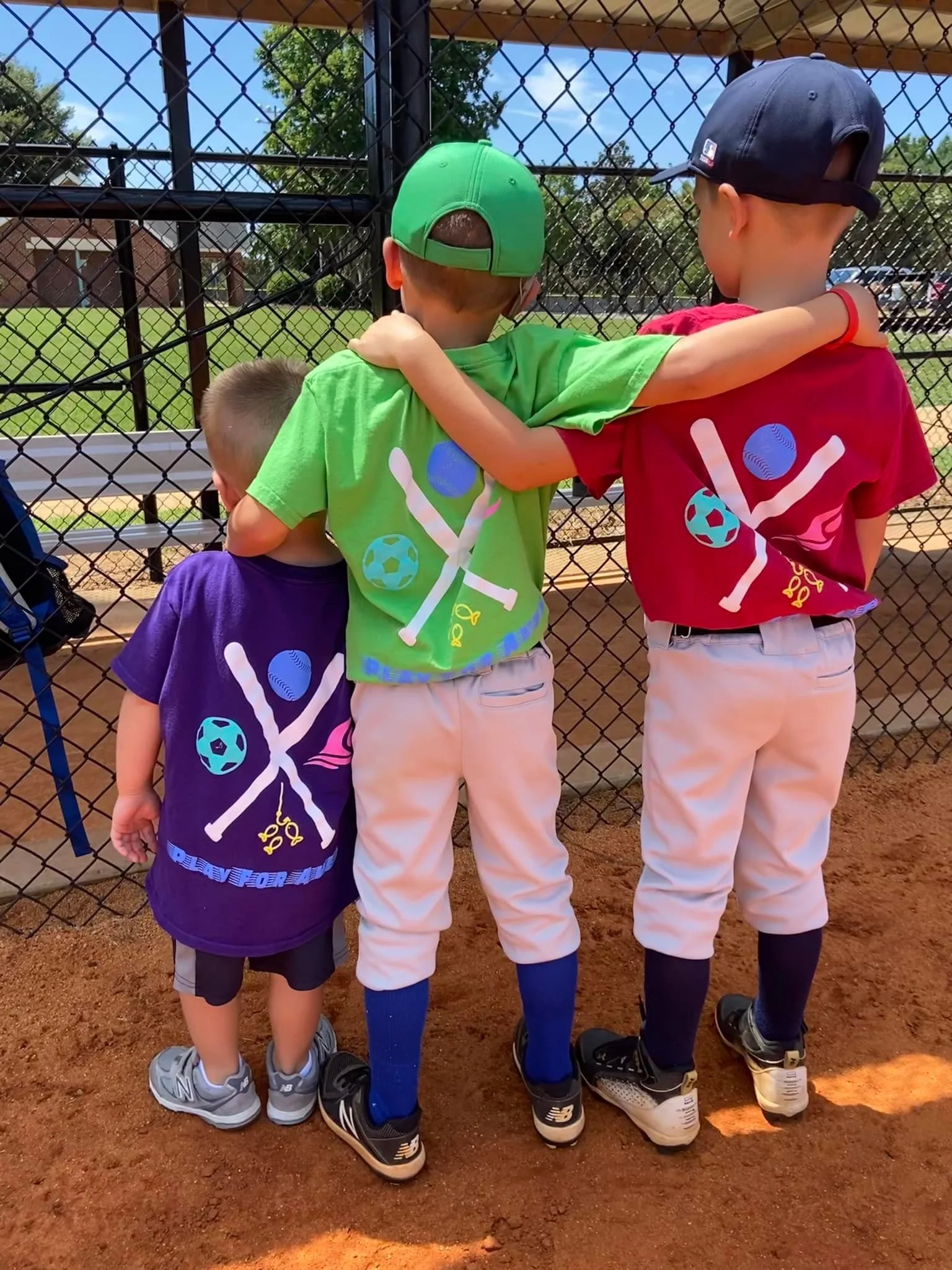 Three young boys standing on a baseball field, facing away from the camera, with arms around each other, looking through a chain-link fence at the game in progress. All are wearing baseball uniforms and sneakers, and two are wearing baseball caps.