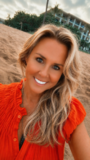 A smiling woman with blonde wavy hair taking a selfie outdoors on a beach, wearing an orange top with ruffled sleeves.