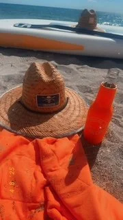 Beach scene with a straw hat, orange towel, and orange drink, with a person lying on a paddleboard in the background.