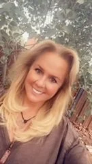 A woman with blonde hair smiling outdoors, with trees and a wooden fence in the background.