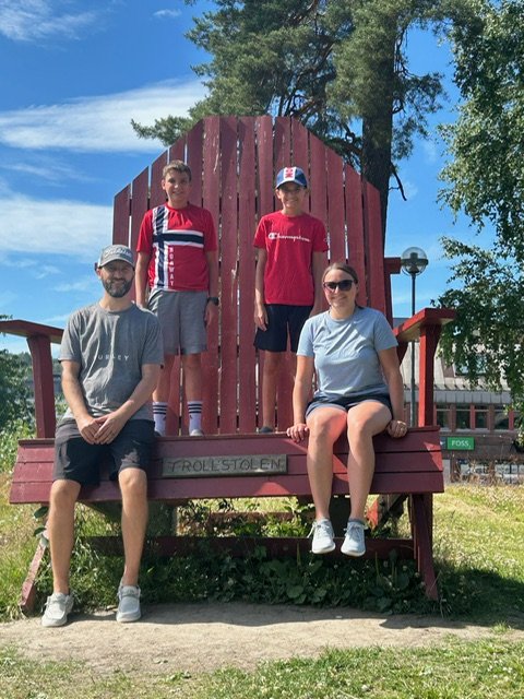 Family of four posing on a giant wooden chair outdoors with trees and a blue sky in the background.
