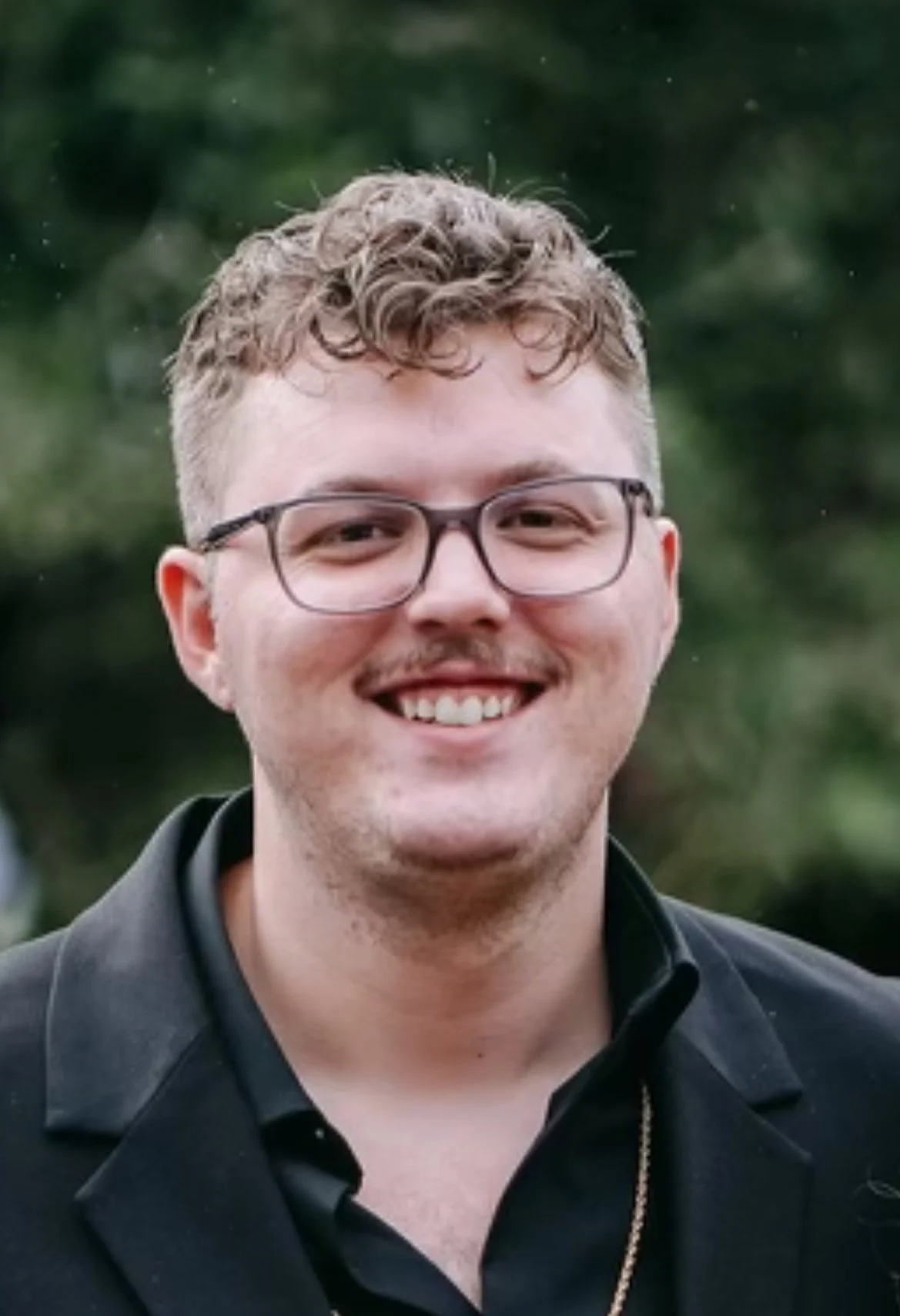 A smiling young man wearing glasses and a black shirt outdoors.