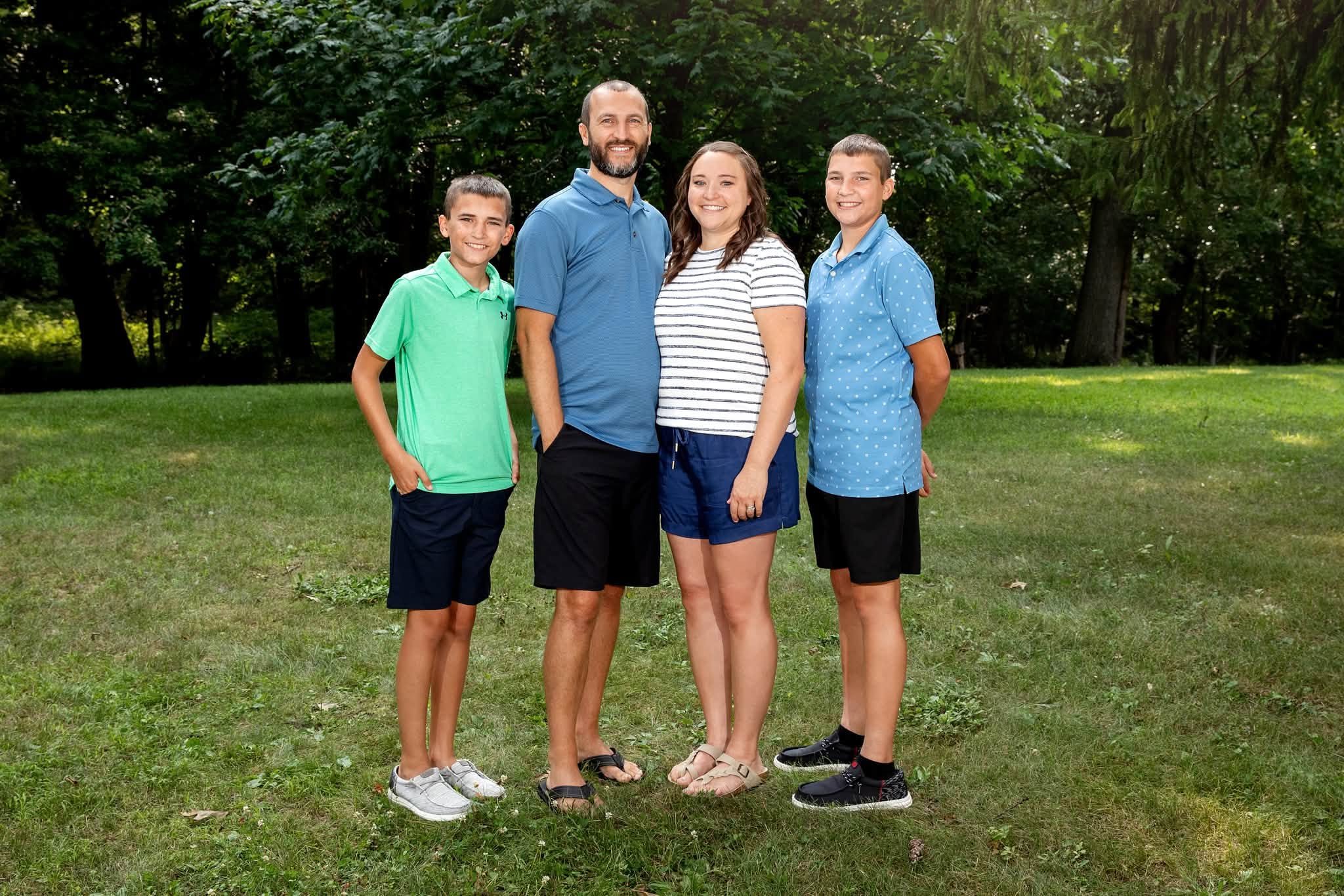 A family of five stands on grass in a park with green trees in the background, smiling at the camera. The group includes two adult adults and three children, all dressed casually.