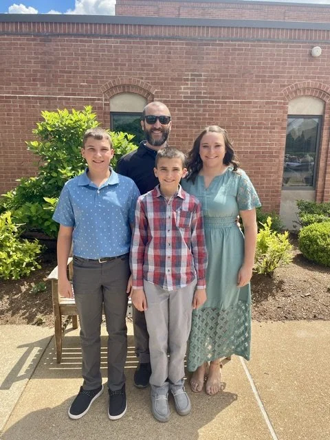 A man and three children standing outside in front of a brick building with green bushes. The man has a beard and wears sunglasses. The children are two boys and a girl, all smiling and dressed in casual clothing.