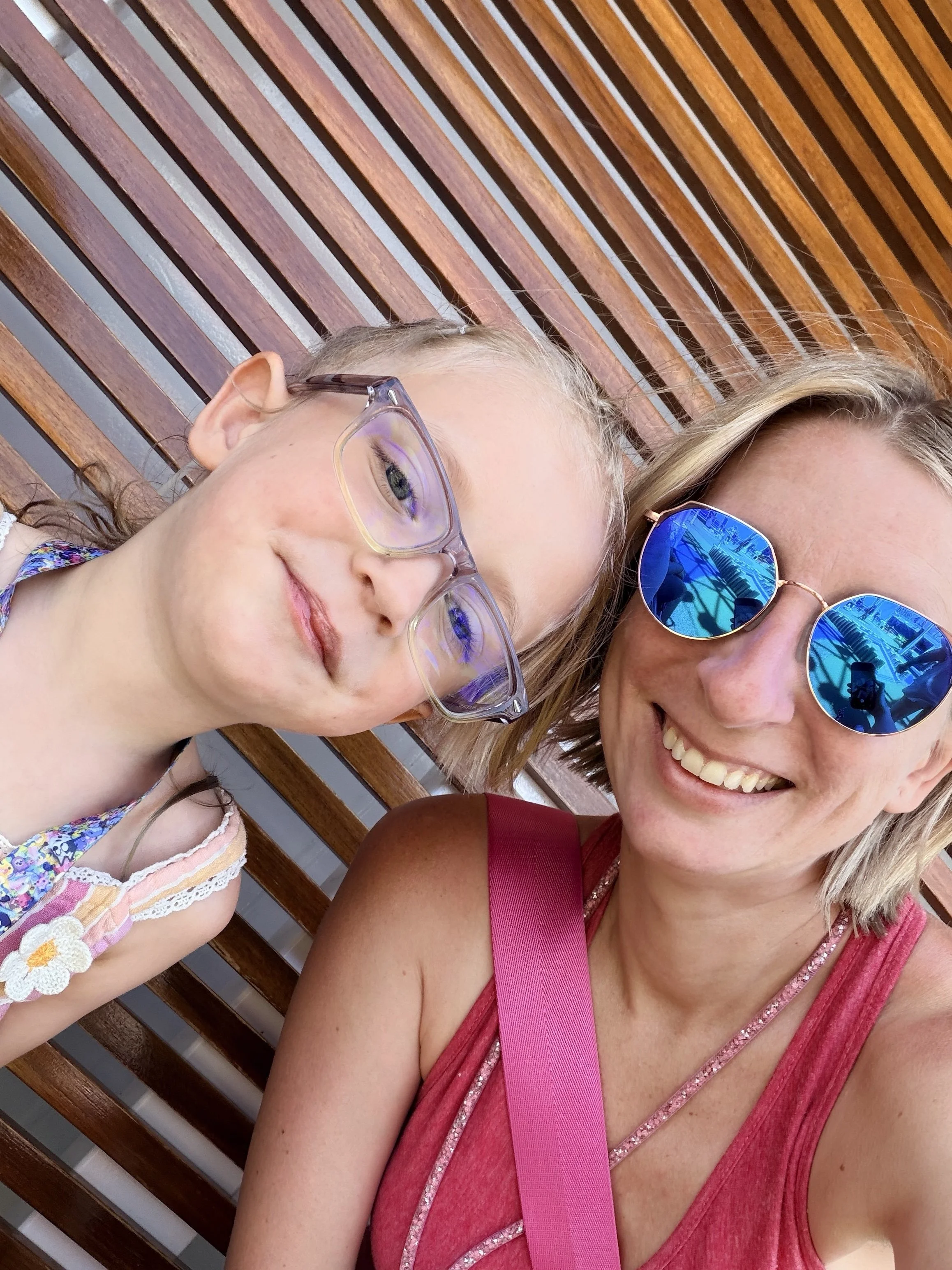 A woman and a young girl taking a selfie together, both wearing sunglasses, sitting on a wooden bench against a background of wooden slats.