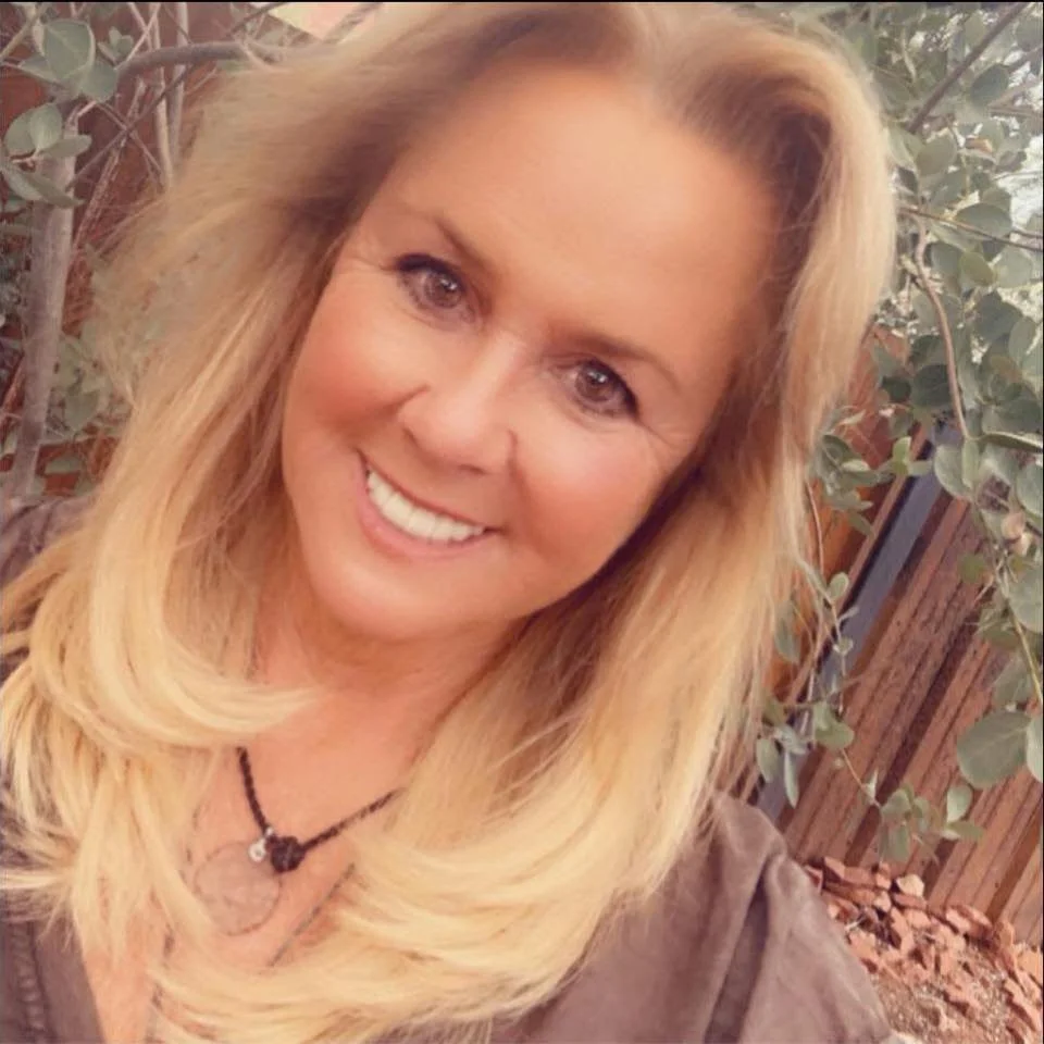 A woman with blonde hair smiling outdoors, with trees and a wooden fence in the background.