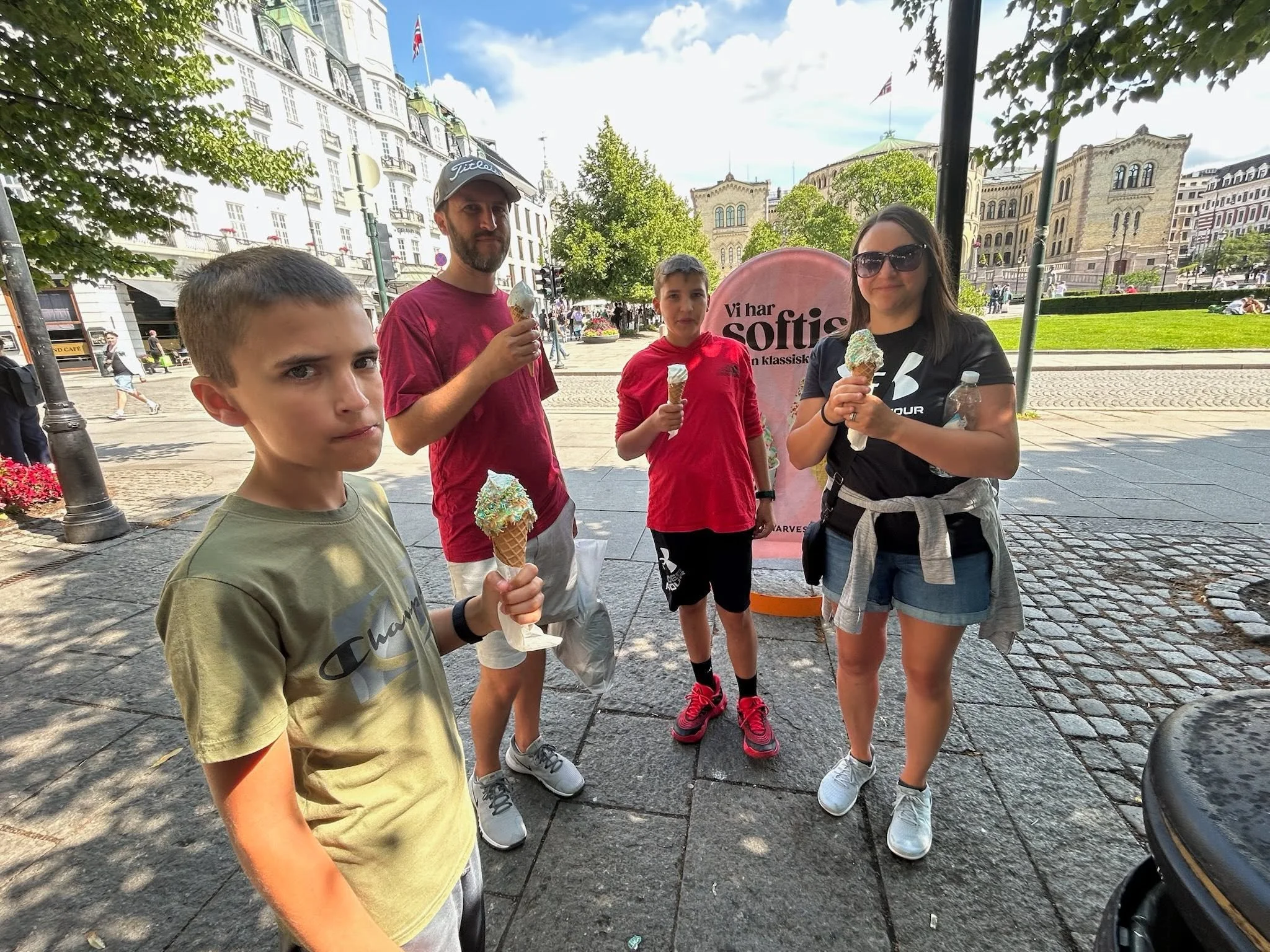 Four people standing outdoors on a cobblestone street, holding ice cream cones, with historical architecture and trees in the background.