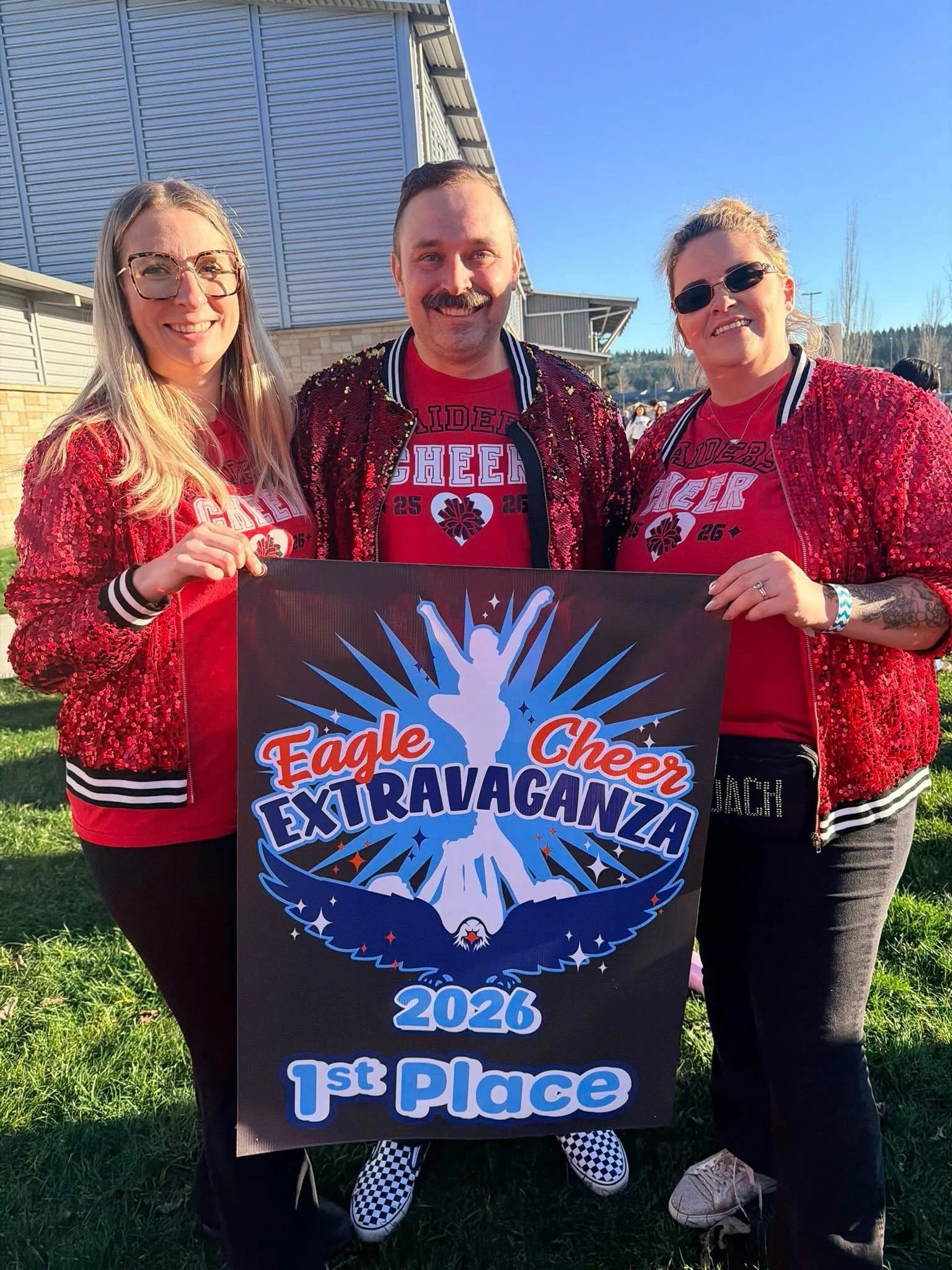 A group of three people outdoors holding a large sign that reads 'Eagle Cheer Extravaganza 2026 1st Place.' They are dressed in red sequined jackets and red shirts, smiling. The person in the middle has a mustache, and the woman on the right is weari