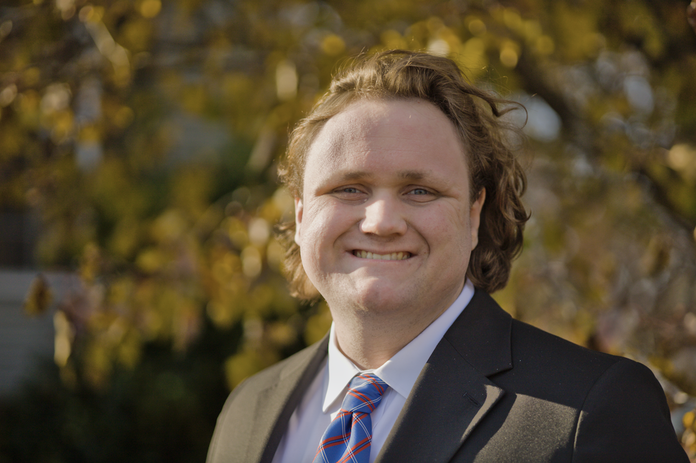 headshot of man in suit outdoors with golden leaves in background