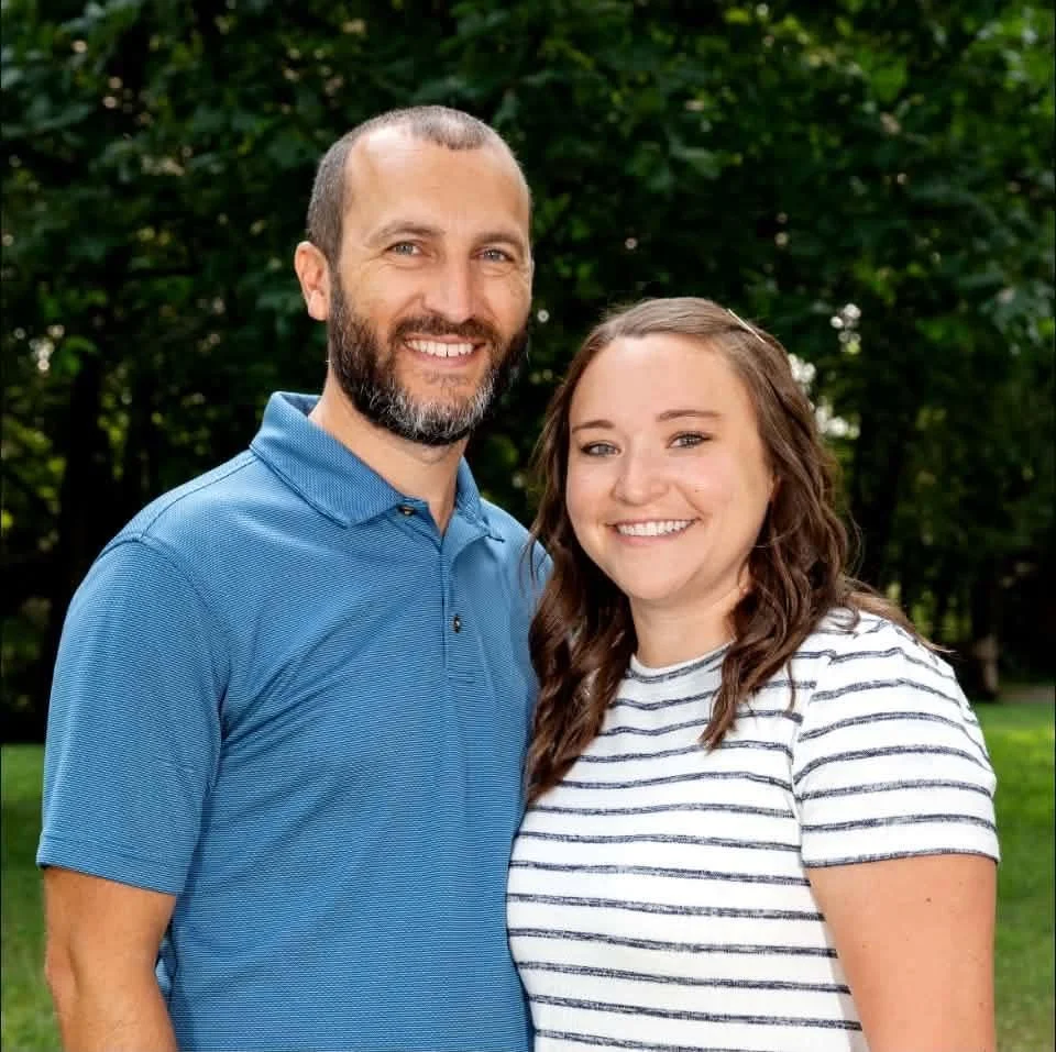 A man with a beard and a woman with long brown hair smiling outdoors, standing close together in front of green trees.