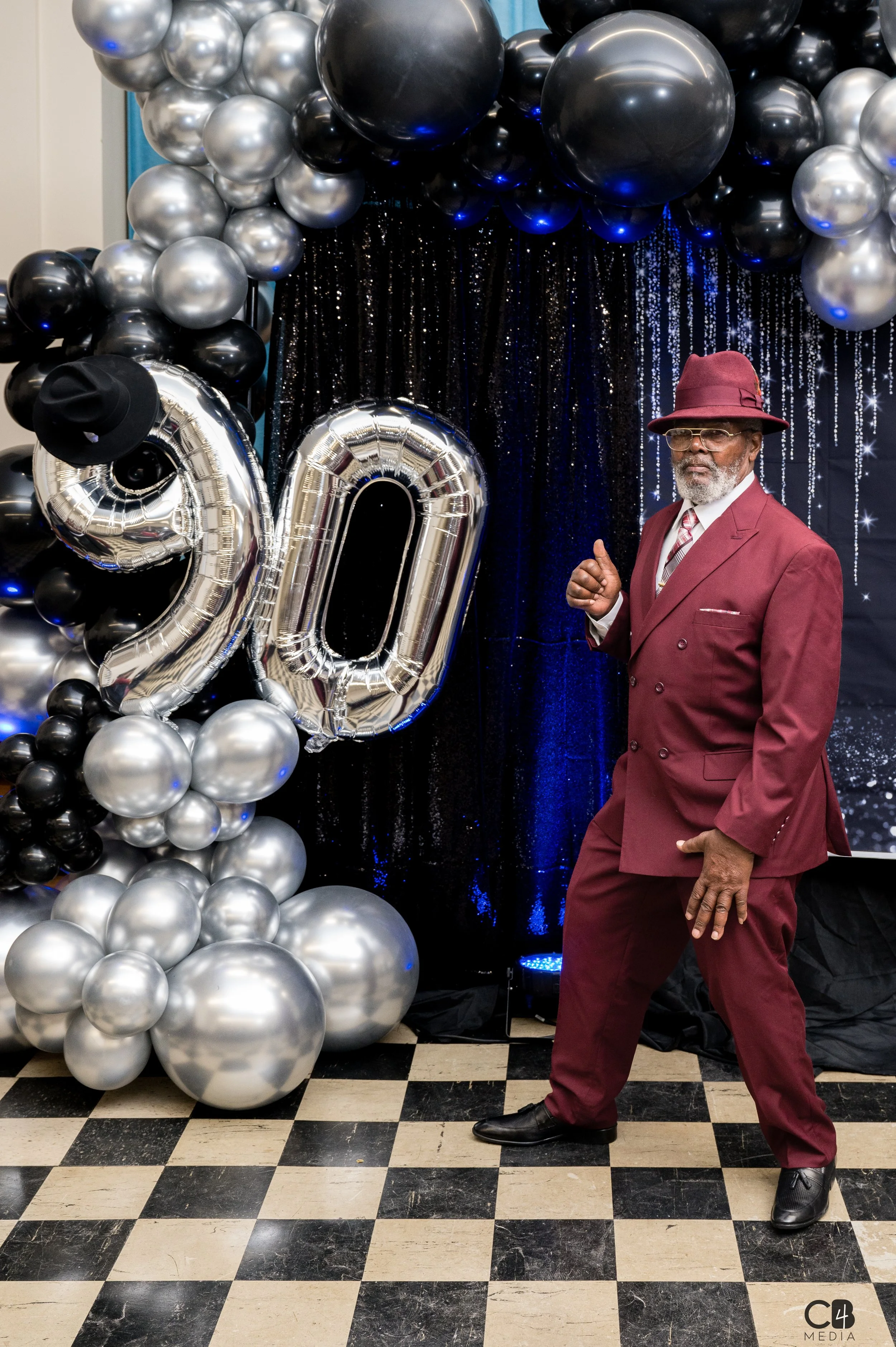 A man in a burgundy suit and hat standing on a checkered floor at a Birthday celebration with black, silver, and blue balloons and large silver balloons forming the number 90.